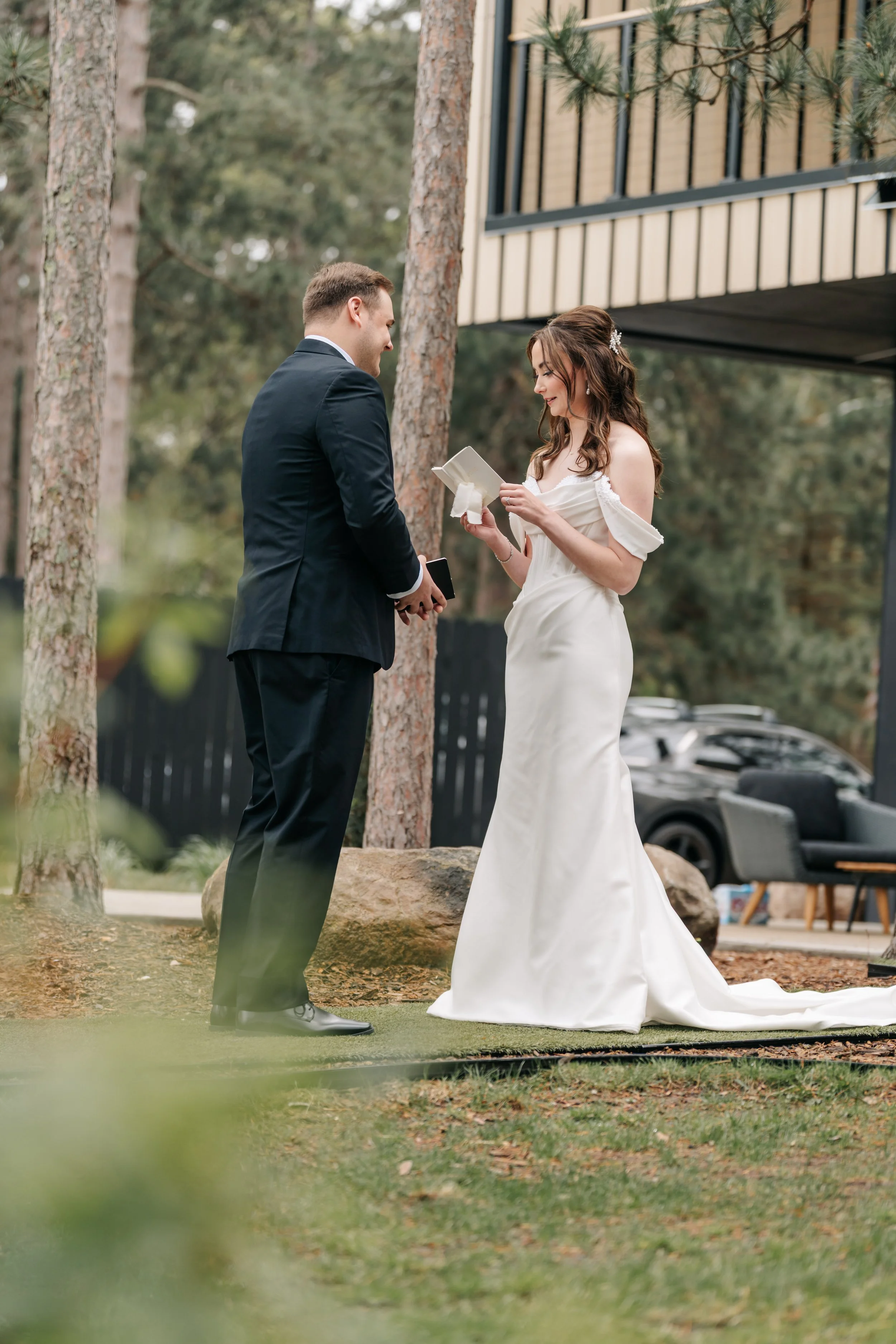 A bride and groom exchanging vows outdoors during daytime, surrounded by trees and rocks, with a building and cars in the background.