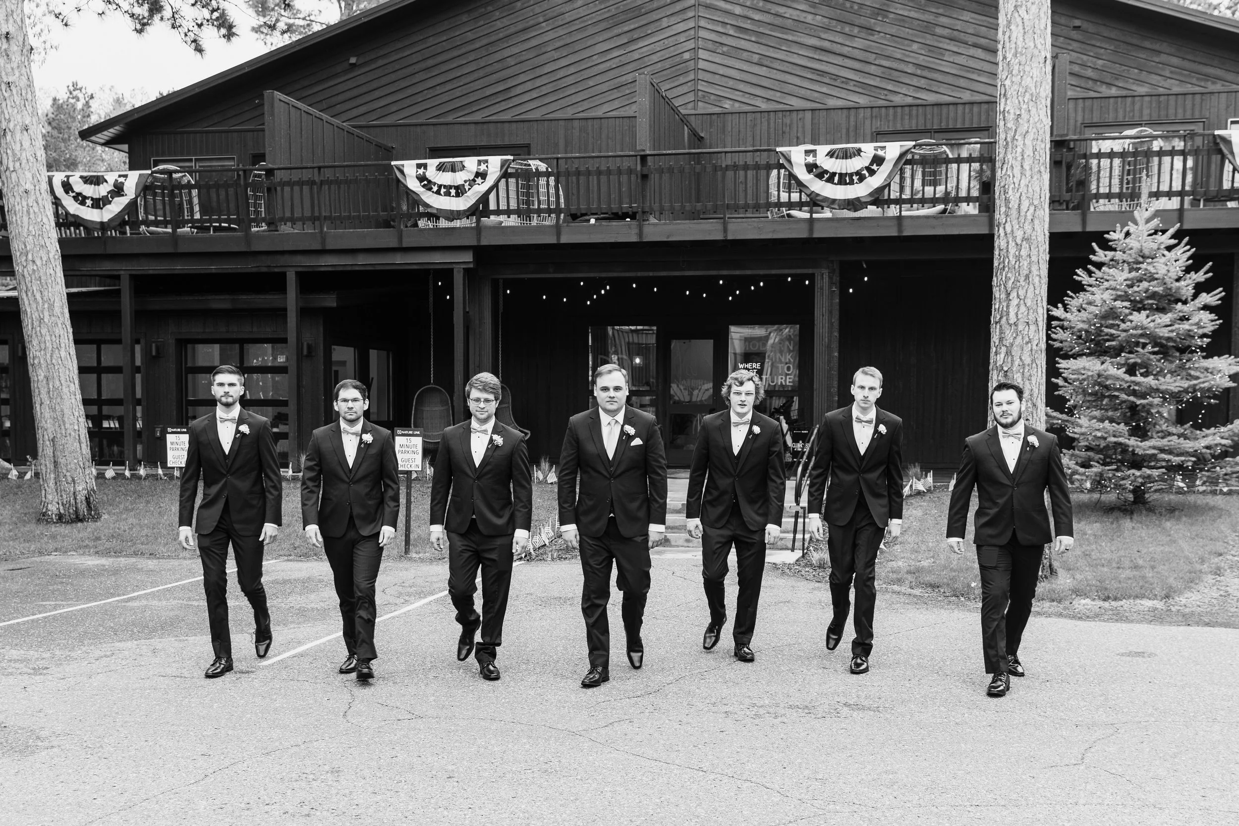Seven men in tuxedos walking in a line in front of a building decorated with patriotic bunting, with trees and a Christmas tree on the right.