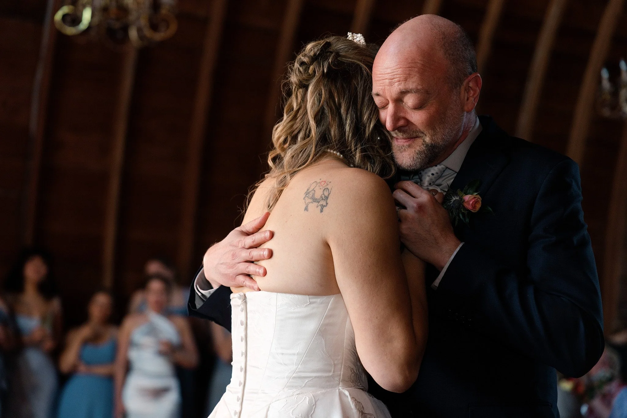 A bride and her dad are dancing during their wedding reception in a decorated venue, with guests watching in the background.