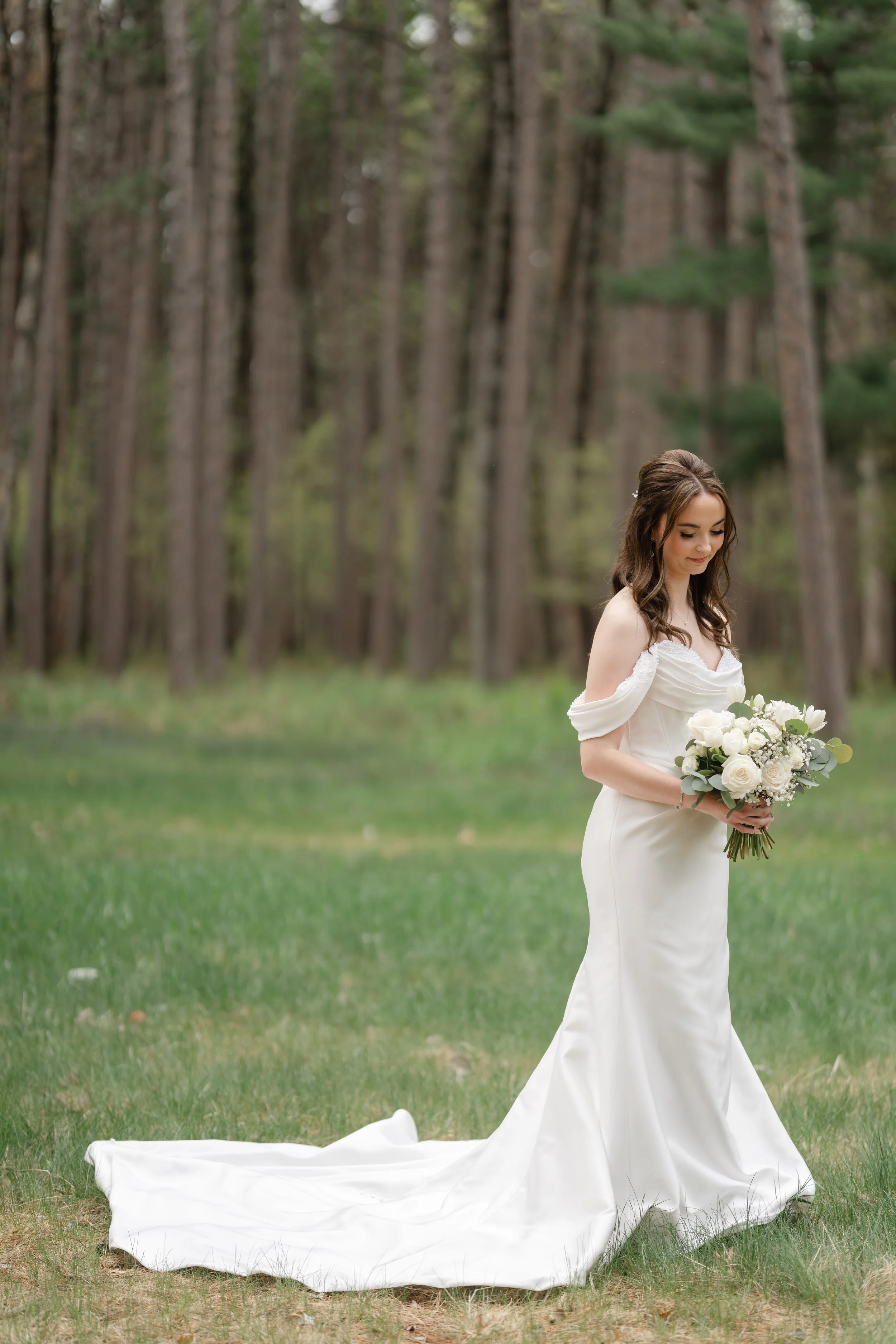 A bride in a white wedding dress holding a bouquet of white flowers standing on grass in a forested area.