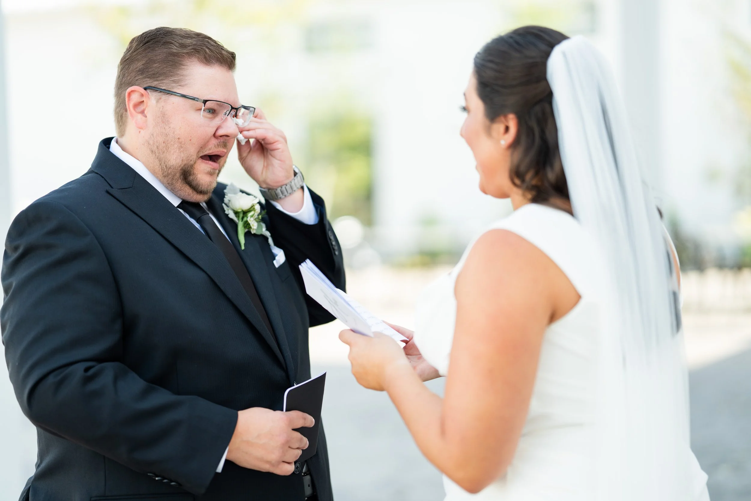 A bride and groom during their wedding ceremony outdoors, with the bride reading vows and the emotional groom, holding a small book, both dressed in formal wedding attire, taken by MN photographer LKW Photography.
