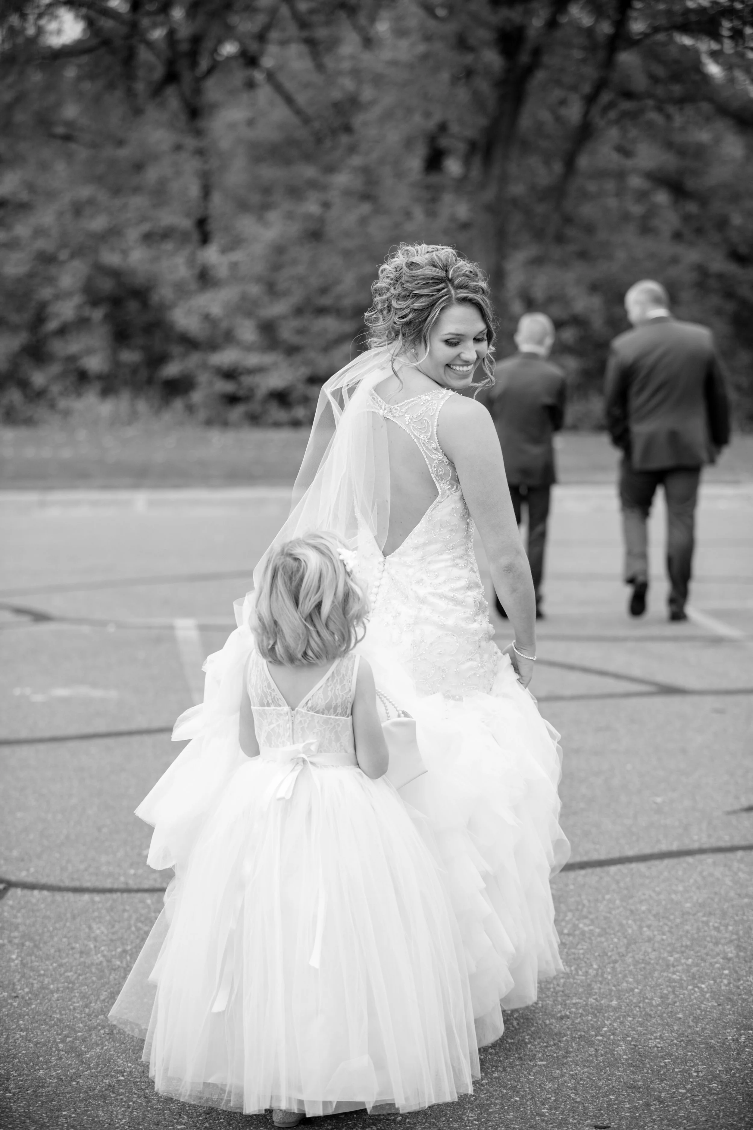 Minnesota based photographer, LKW Photography, photographs a bride in a wedding dress smiling and looking back at a young girl in a white dress with a bow, who is trying to carry her dress.