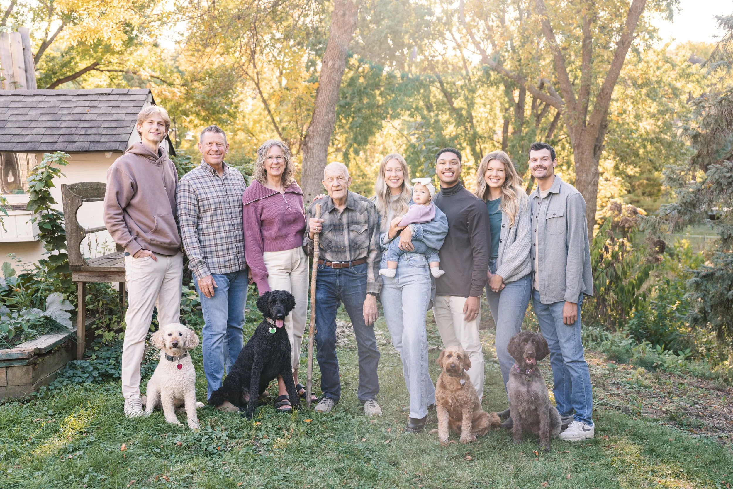 Minnesota portrait photographer, LKW Photography, photographs a family outdoors with multiple generations, including elderly with cane, children, young adults, and dogs, in a backyard with trees and sunlight.
