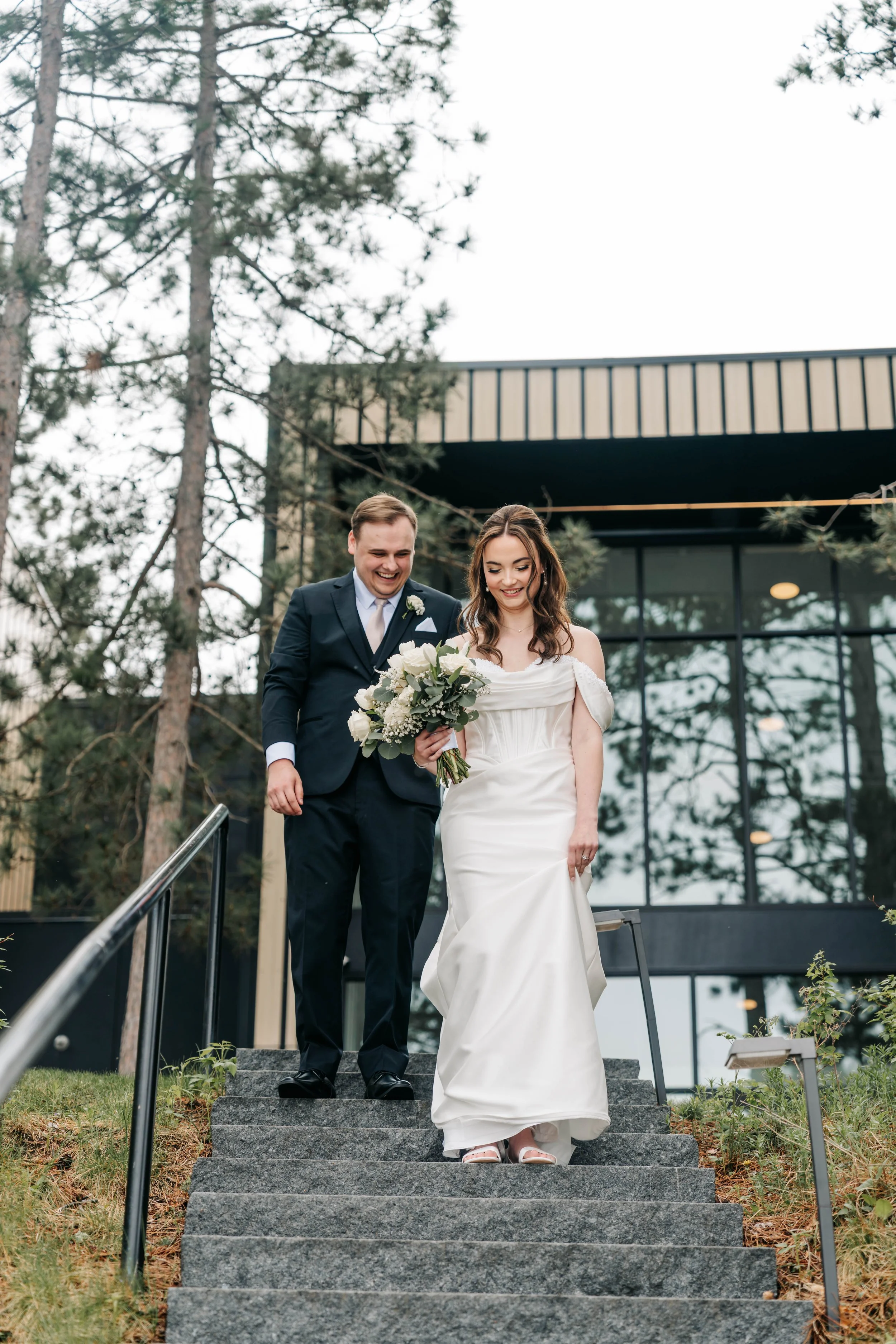 A bride and groom are descending outdoor stone stairs, with the bride holding a bouquet of white flowers, both smiling, against the backdrop of a modern building and tall pine trees.