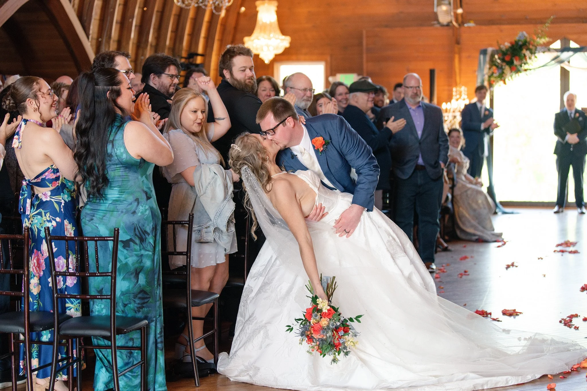 Bride and groom just married and stop halfway down the aisle for a beautiful dip kiss in front of their guests.  The wedding takes place in a barn like venue called Green Acres Events.