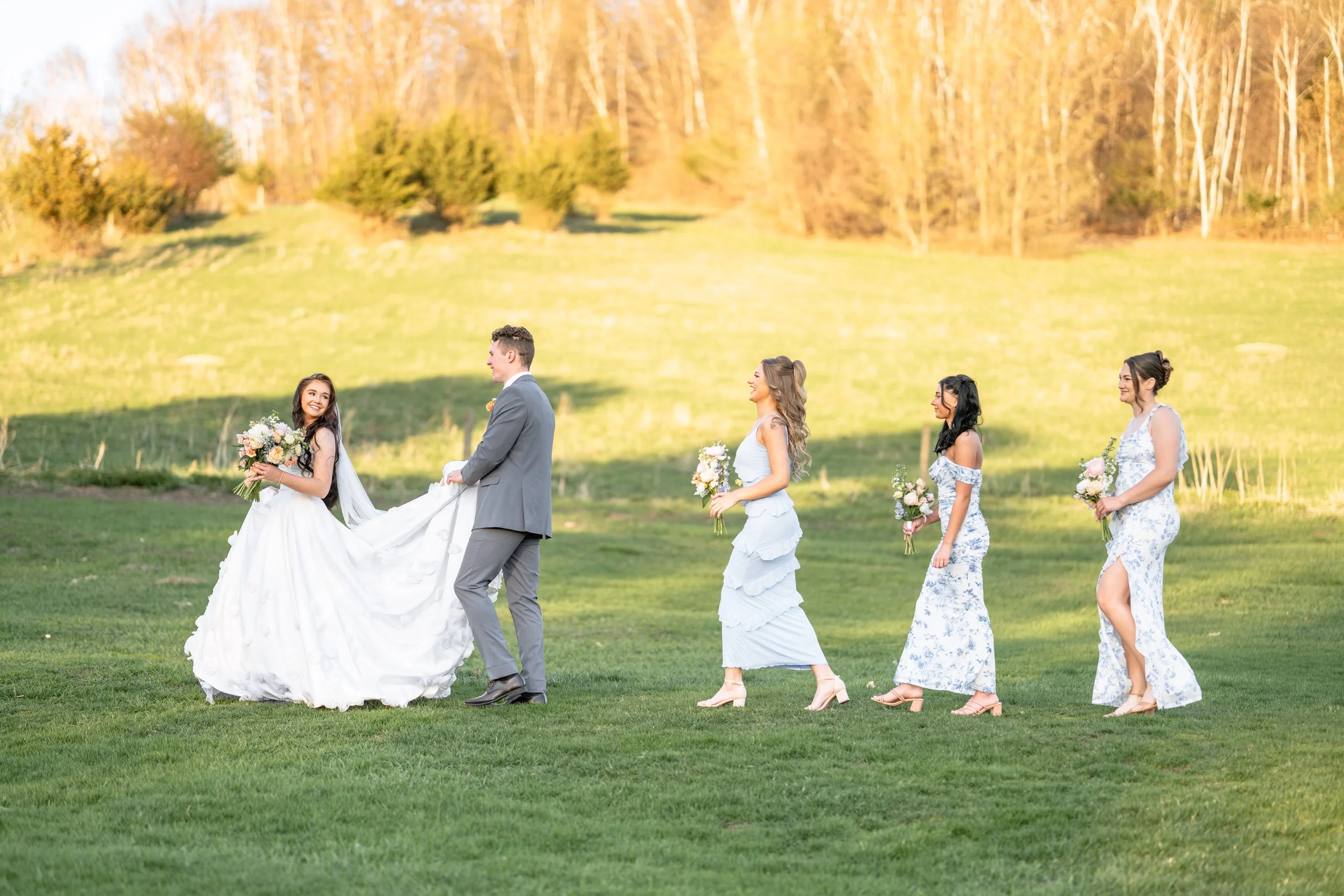 When the wedding is finished and the wedding party is walking from the ceremony site to the reception site at Willow Brooke wedding and event center in Minnesota.  The groom is holding the brides dress as she looks back at him, bridesmaids follow.