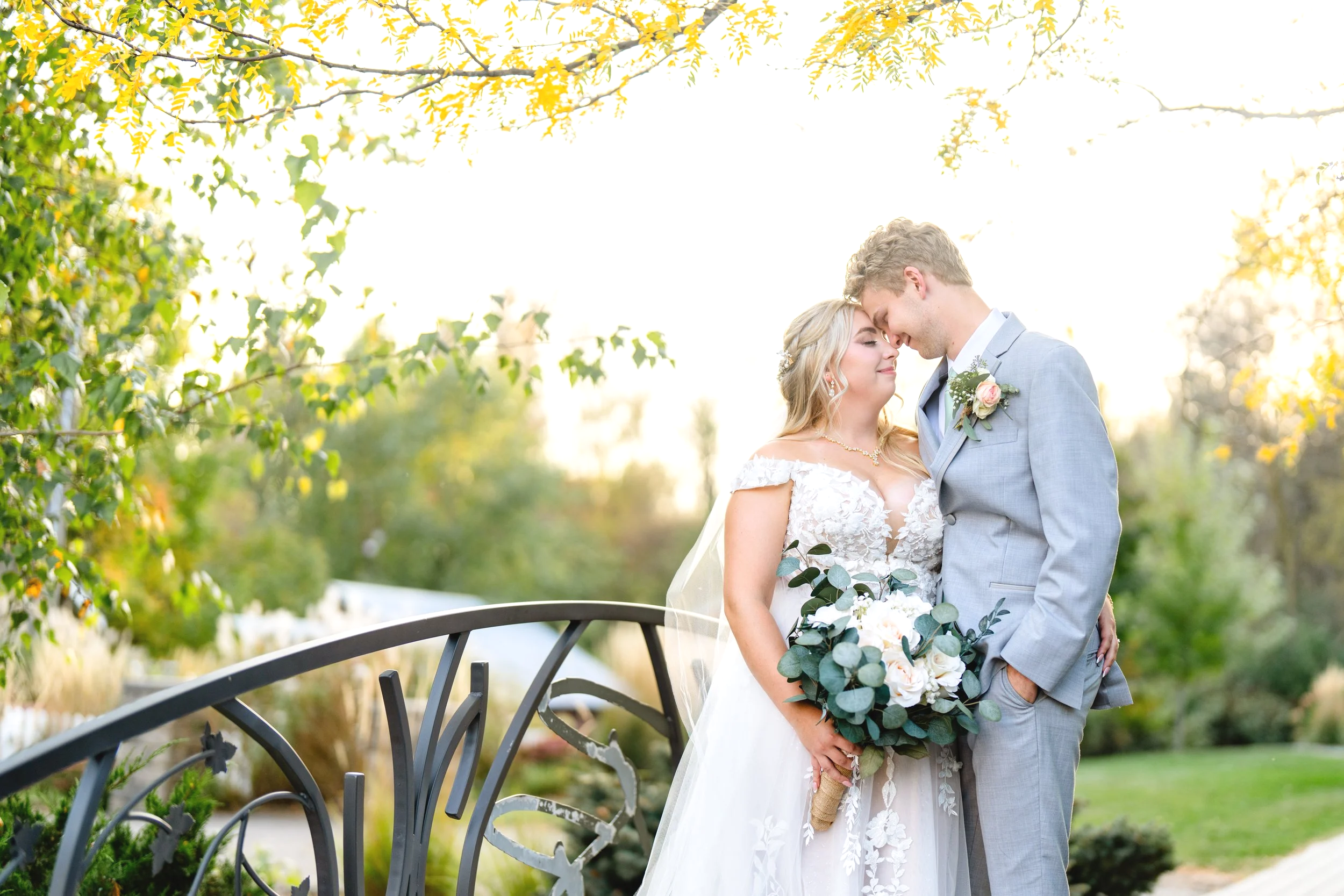 A bride and groom on their wedding day standing close together outdoor with trees and sunlight in the background, the bride holding a bouquet of flowers, both smiling with their faces touching.