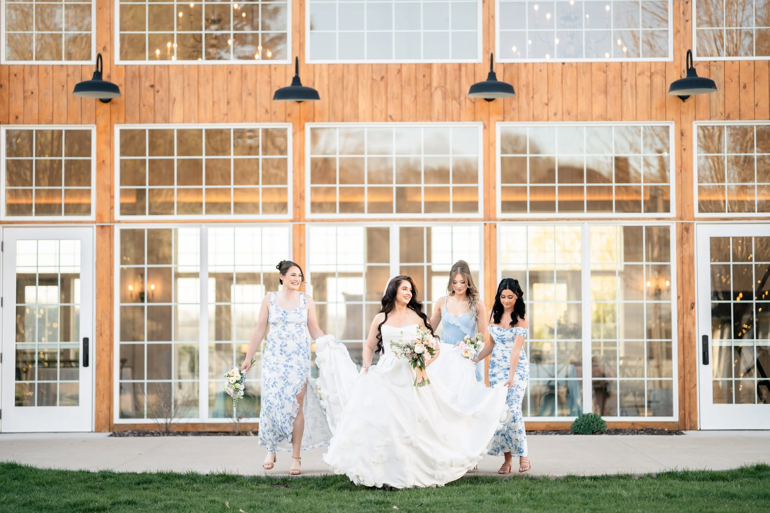 A bride and her bridesmaids are walking, with the bridesmaids holding her dress for her so it doesn't get dirty.  Photographed at Willow Brooke Wedding and Event Center in Minnesota.