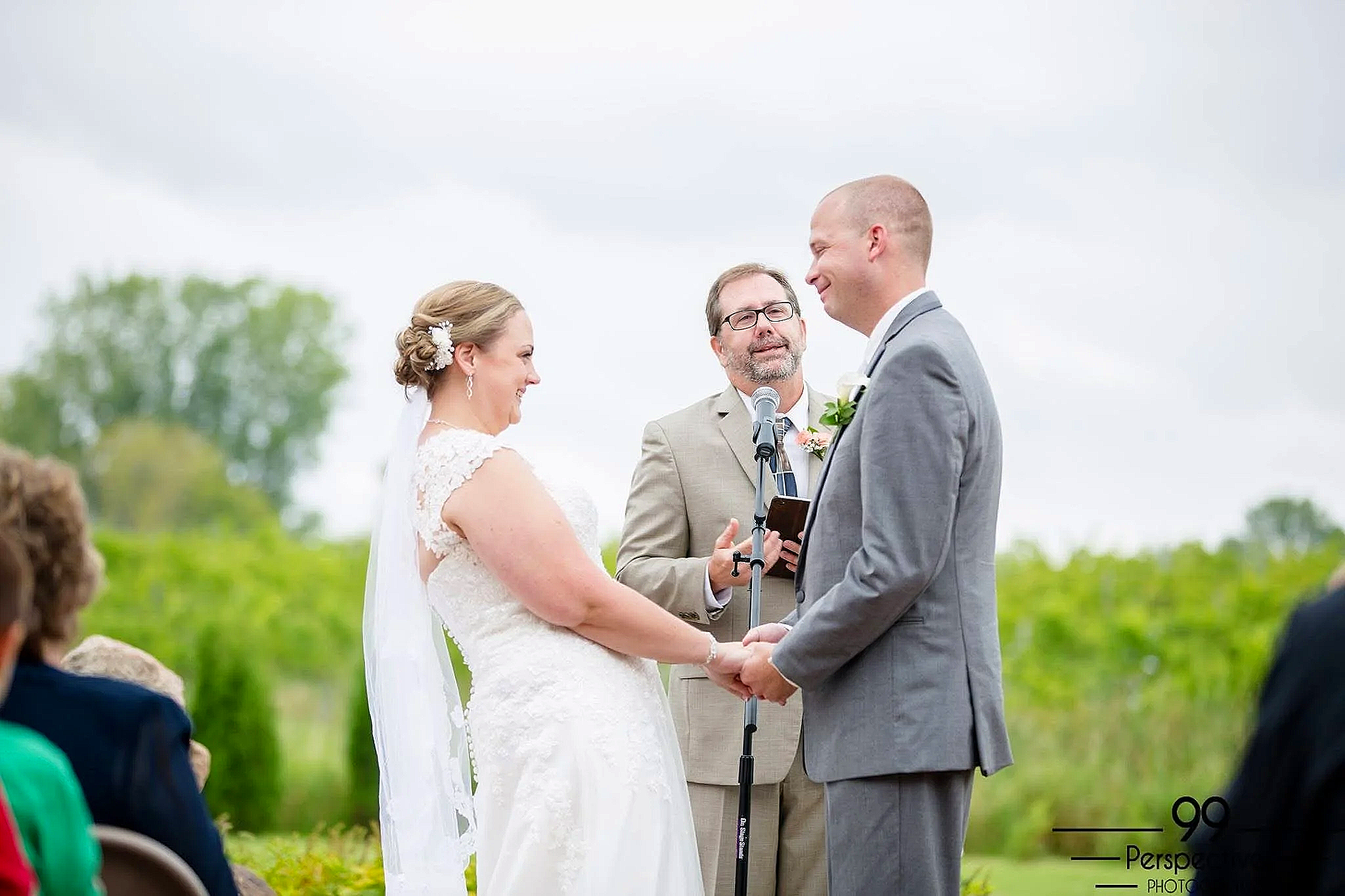 A bride and groom holding hands during their outdoor wedding ceremony with a officiant reading vows, surrounded by friends and family, with green trees and cloudy sky in the background, taken by Minnesota Wedding Photographer LKW Photography.