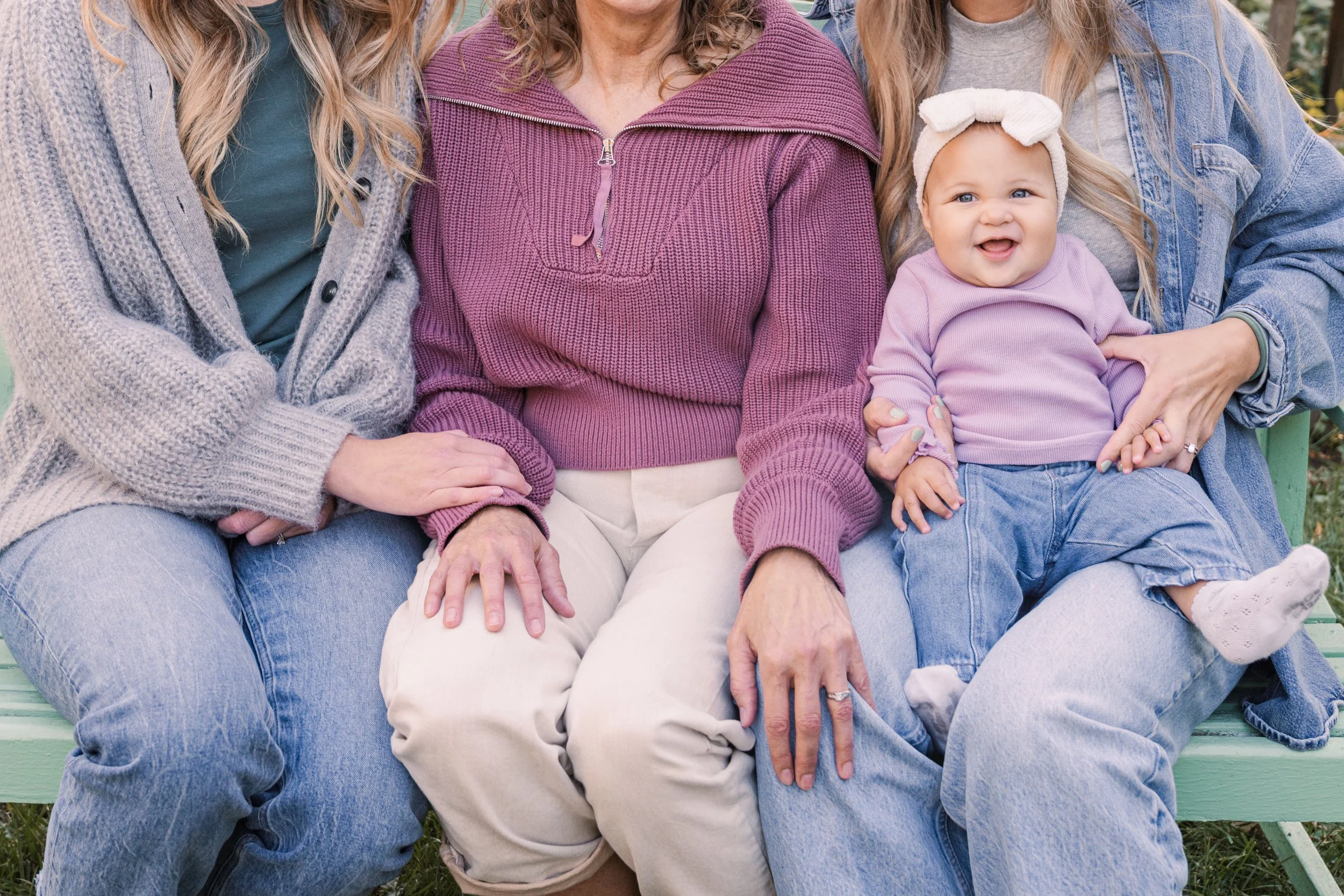 Minnesota family photographer, LKW Photography, photographs a family of a mom, 2 sisters, and a baby, sitting on a green park bench, dressed casually. The baby is smiling and wearing a lavender top and a white headband. 