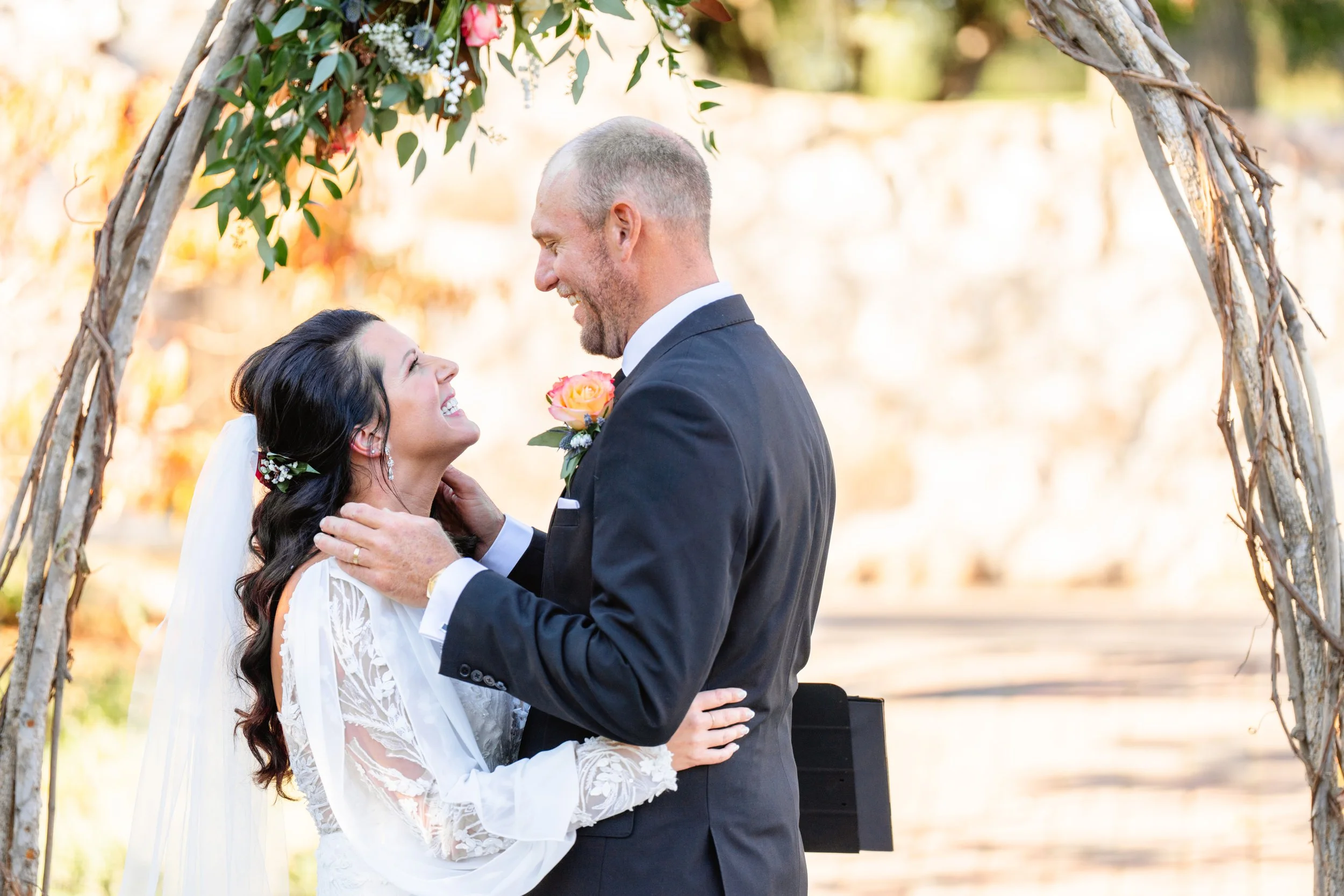 A bride and groom smiling and embracing during their wedding ceremony under a wooden arch decorated with flowers and greenery, taken by LKW Photography based in Minnesota.