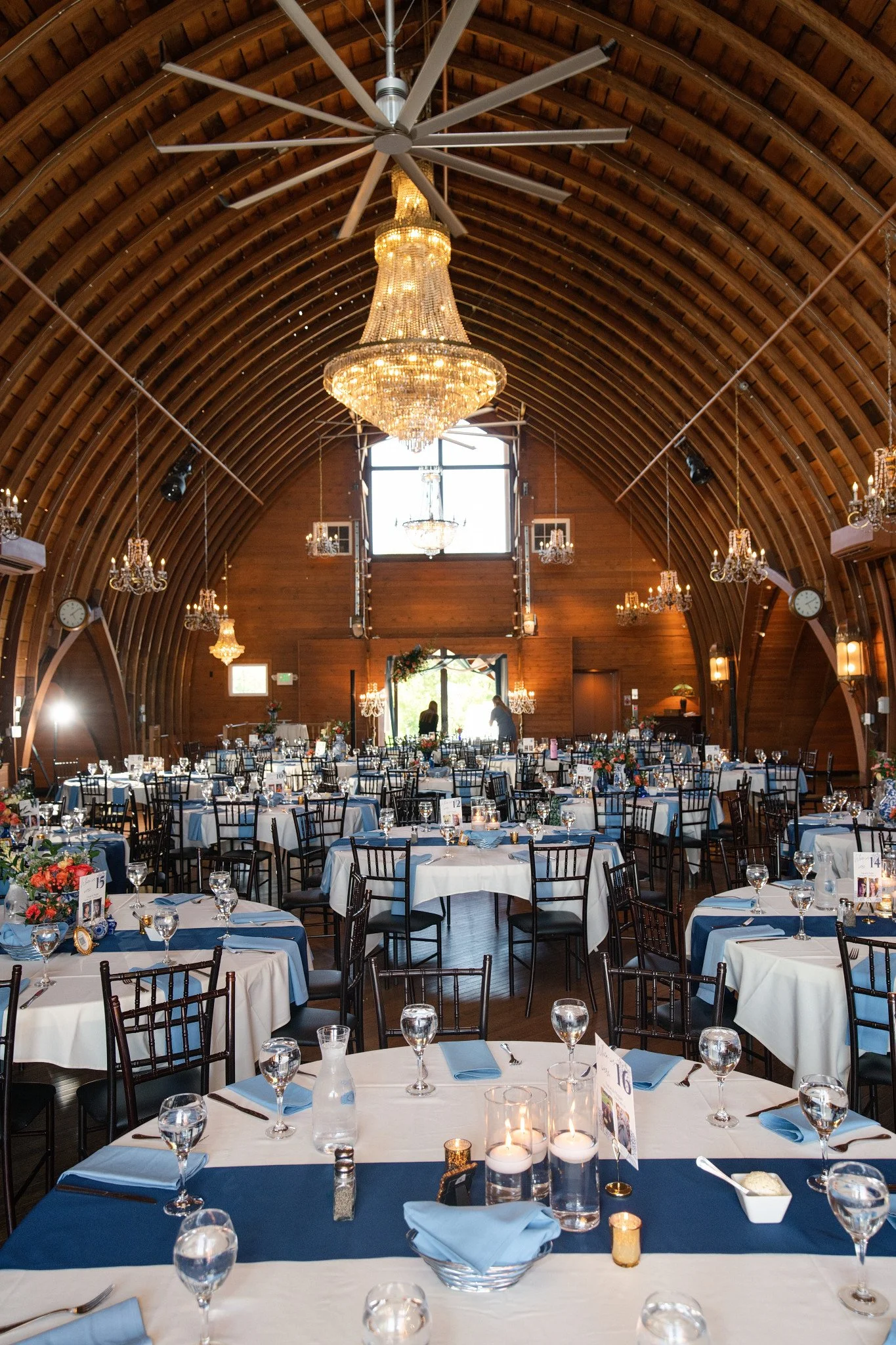 The indoor reception space at Green Acres Events, showcasing the beautiful tablescapes in white, navy, and sky blue, along with blue and white china, beautiful floral arrangements, candles, and a large chandelier hanging above.