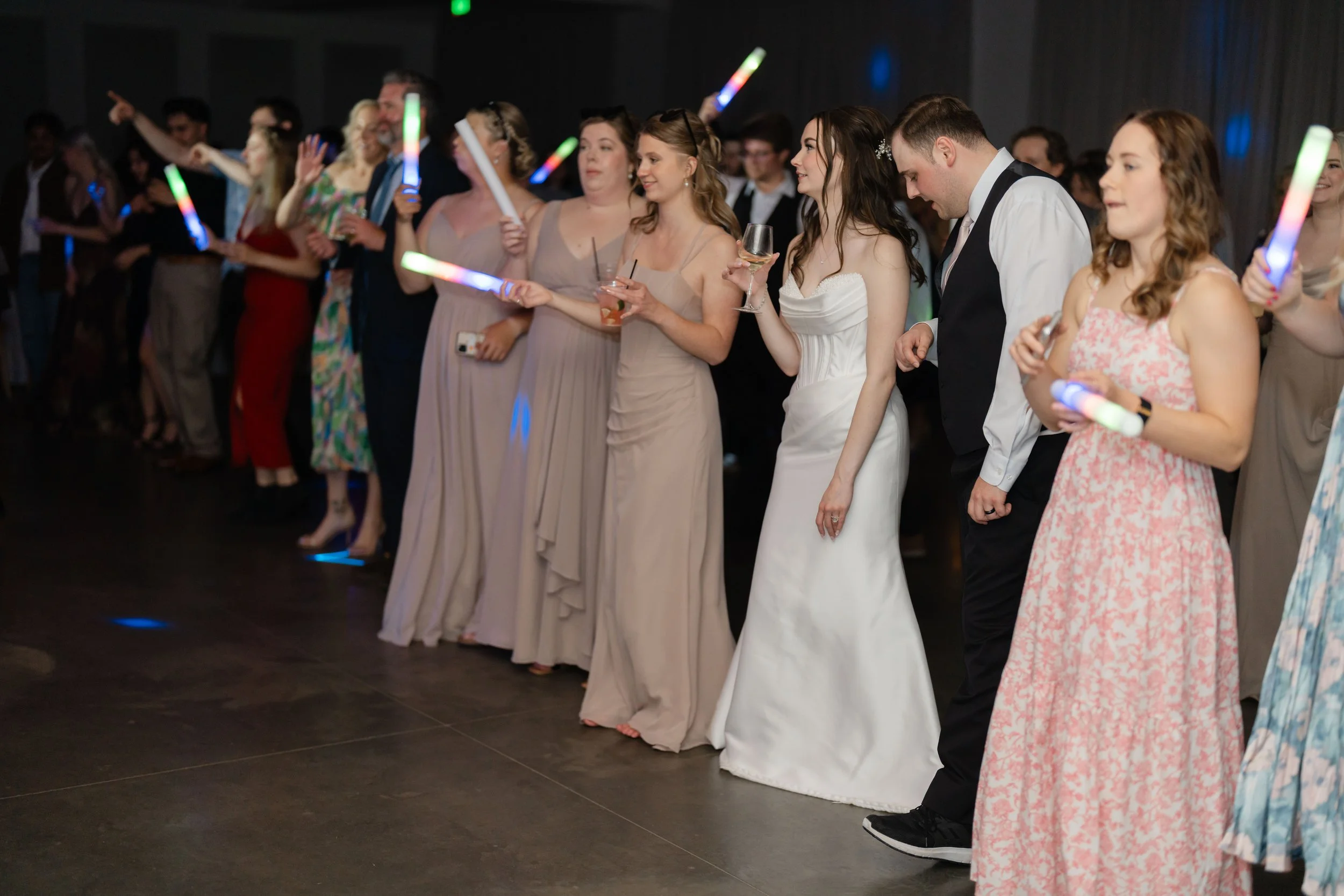 Wedding guests  dancing and holding glow sticks and wine glasses, standing in a line at a celebration or party.