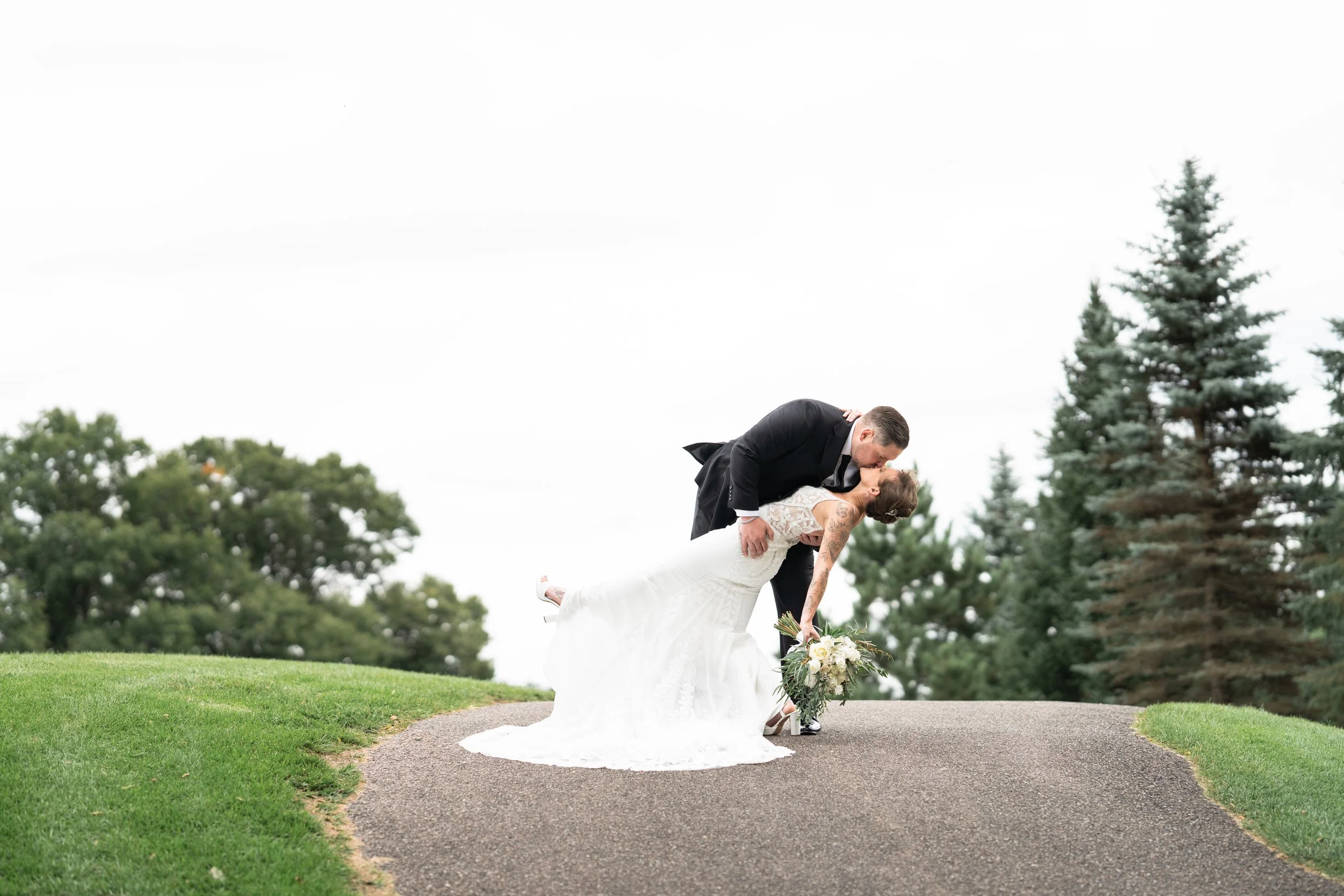 A newlywed couple embraces on a grassy hilltop with trees in the background. The groom, in a black tuxedo, dips the bride in a white wedding dress holding a bouquet of flowers.