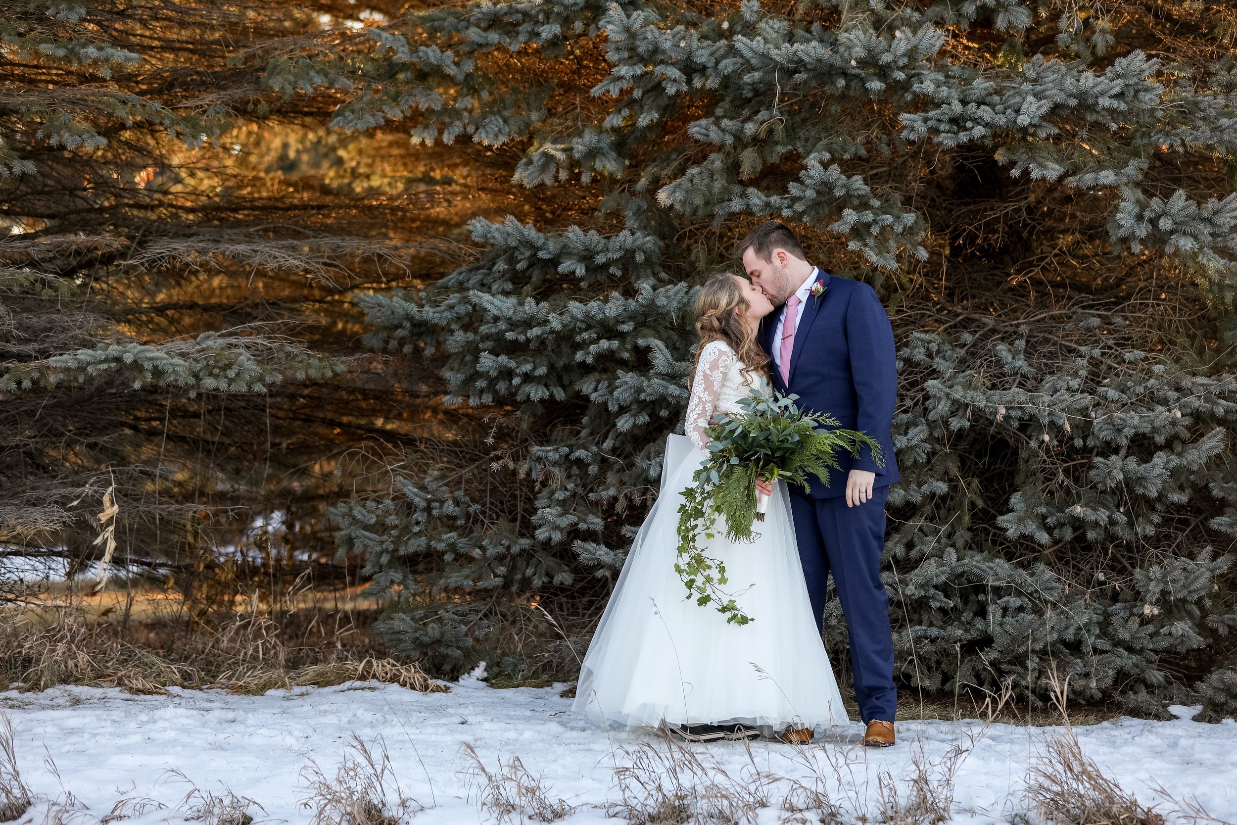 A bride and groom sharing a kiss outdoors in front of a large pine tree, with snow on the ground, taken by MN photographer LKW Photography.