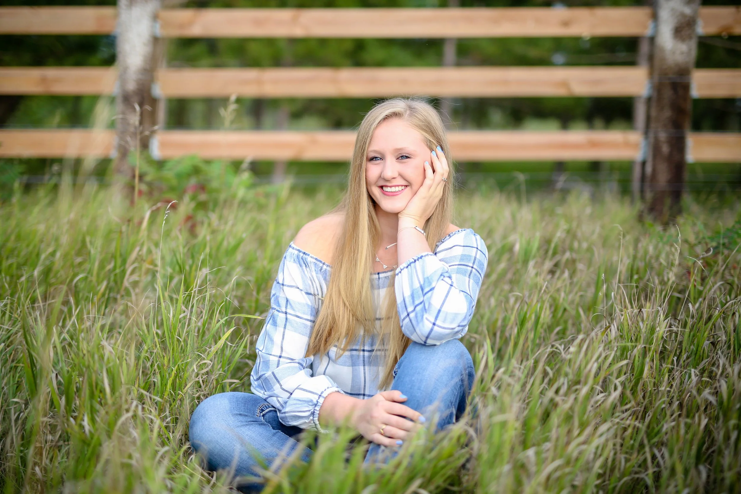 A young woman with long blonde hair sitting on grass in a field, smiling with one hand on her face, wearing a blue and white plaid off-shoulder top and jeans, taken by Minnesota Senior Portrait Photographer LKW Photography.