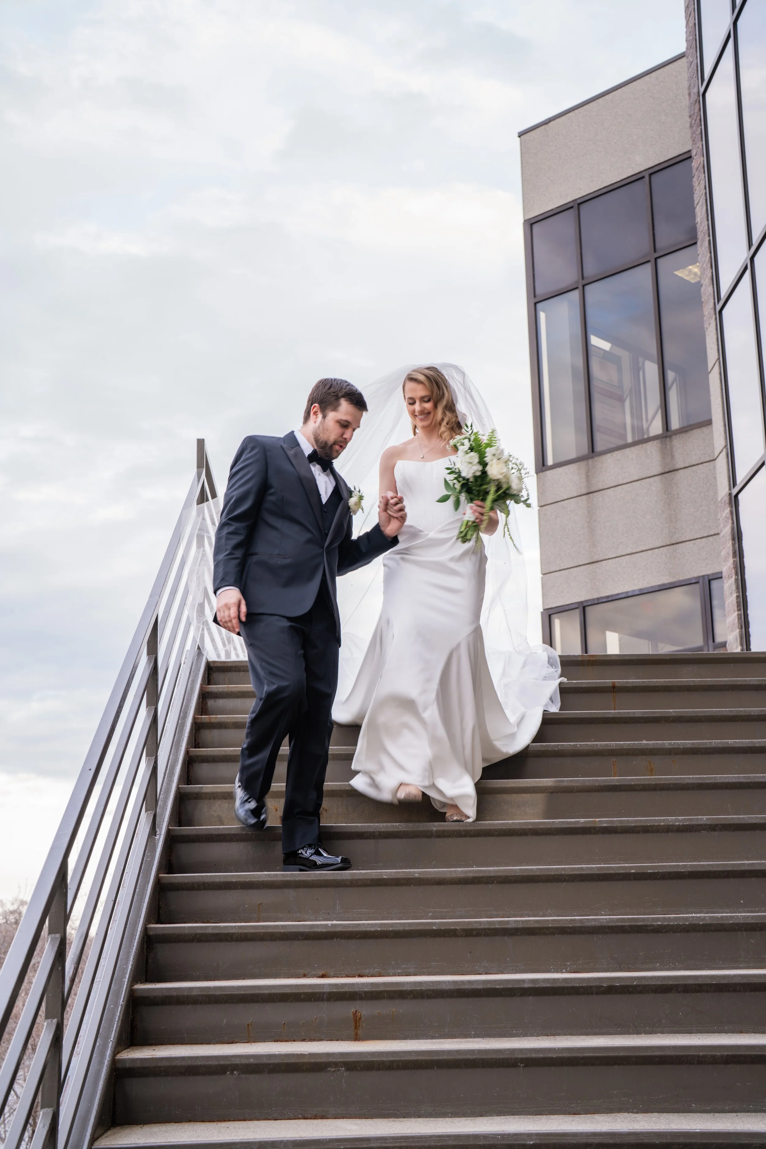 A bride and groom walking down outdoor stairs, holding hands, with the groom in a tuxedo and the bride in a white wedding gown holding a bouquet of white flowers, near a modern building with large glass windows.