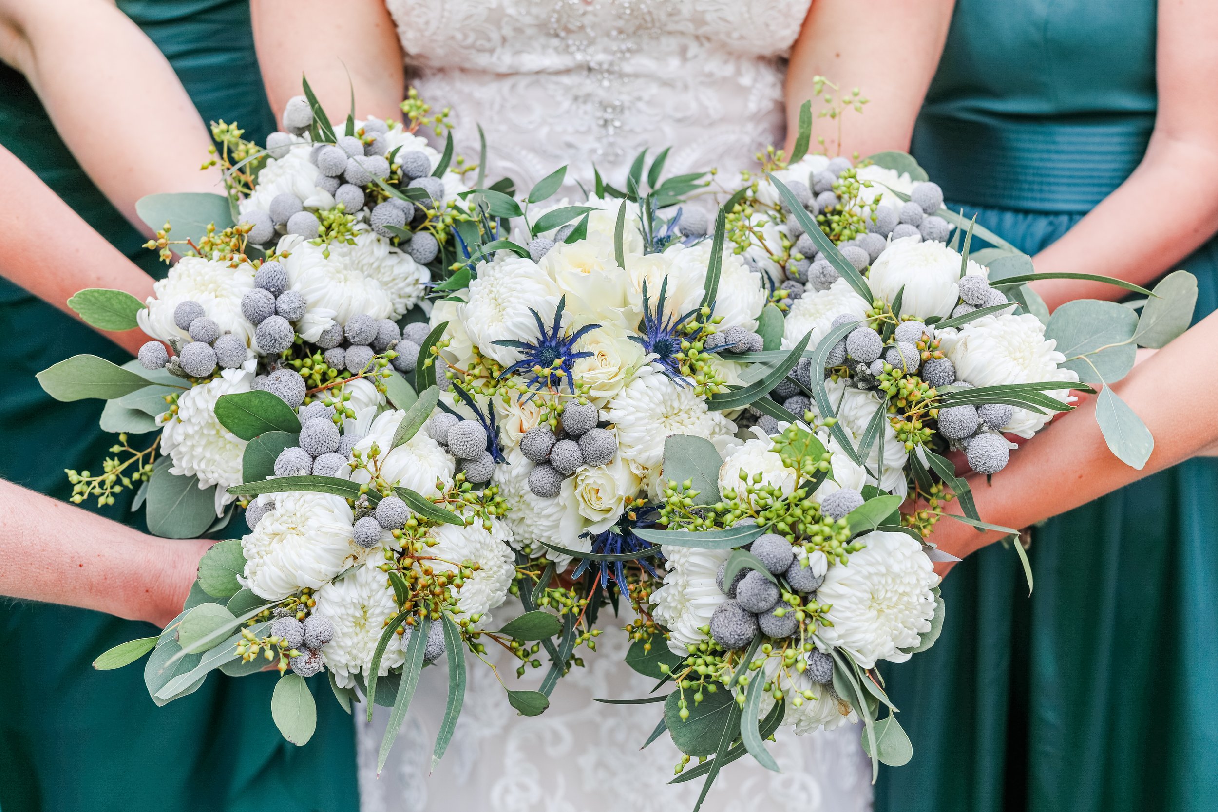 Bride holding a bouquet of white flowers with green leaves and purple accents, flanked by bridesmaids in green dresses, photographed by Minnesota based wedding photographer, LKW Photography.