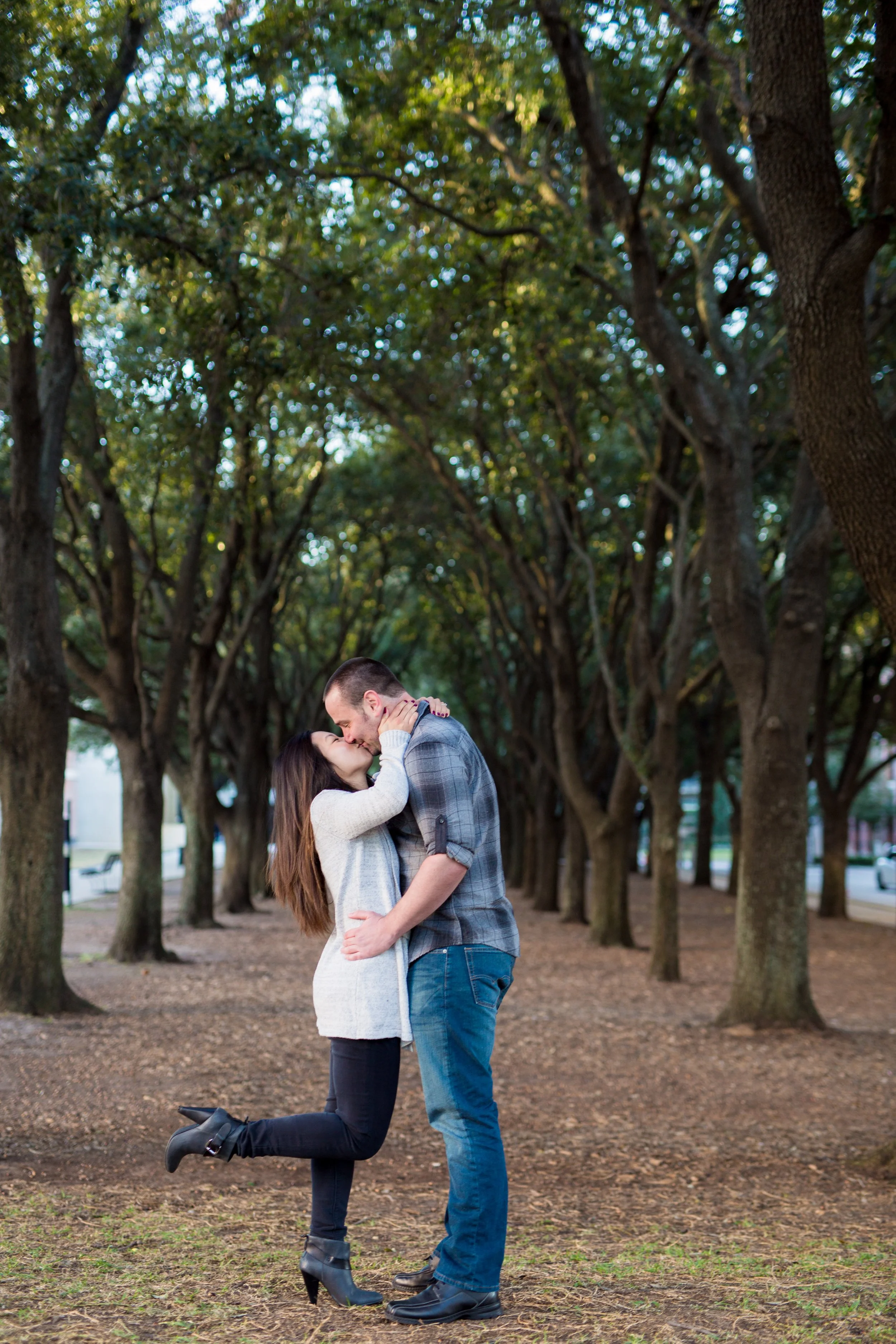 A couple kissing under a row of tall trees in a park, taken by MN photographer LKW Photography, in Austin, TX.