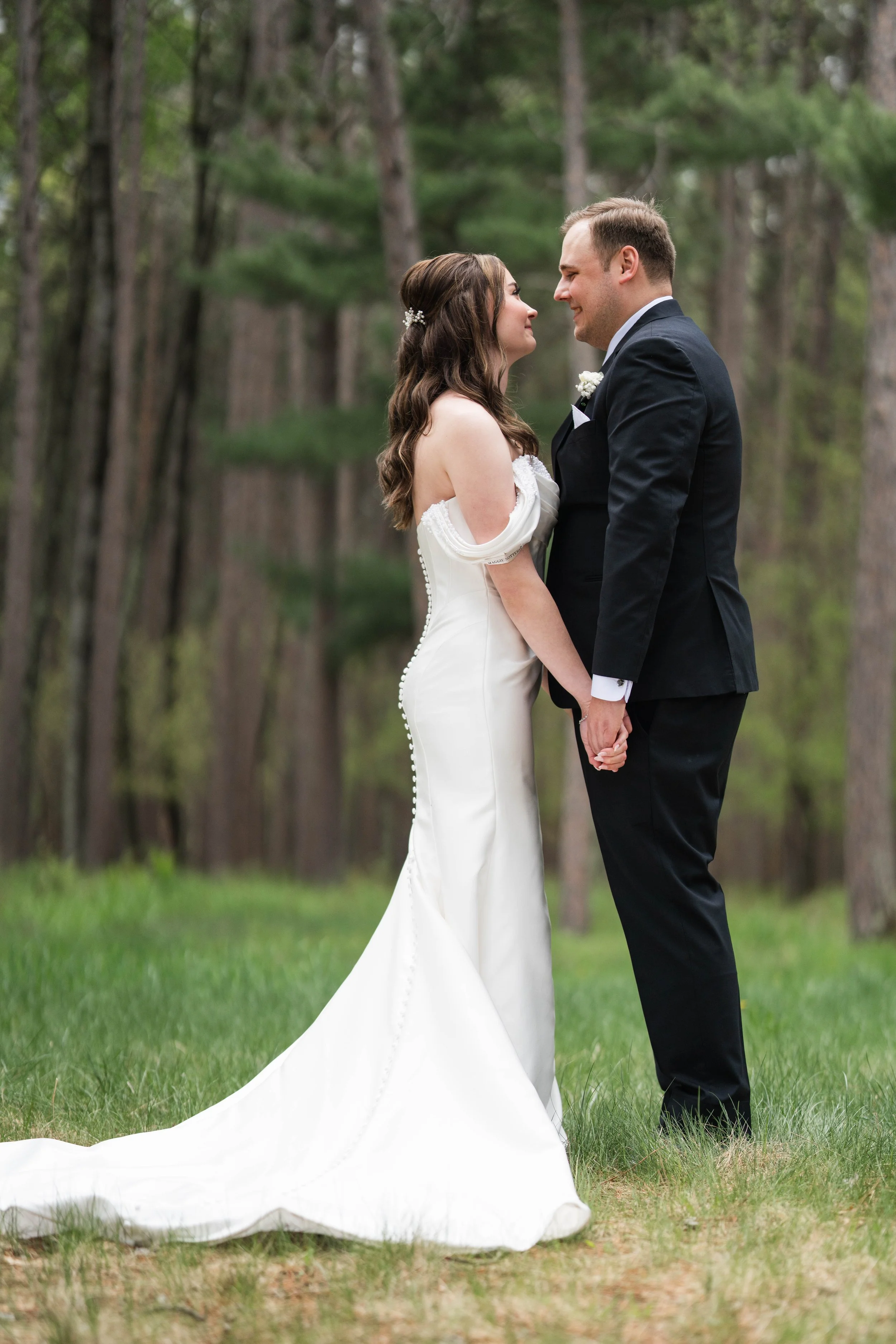 A bride and groom standing close together outdoors among trees, holding hands and gazing at each other, with the bride wearing a white wedding dress and the groom in a black suit.