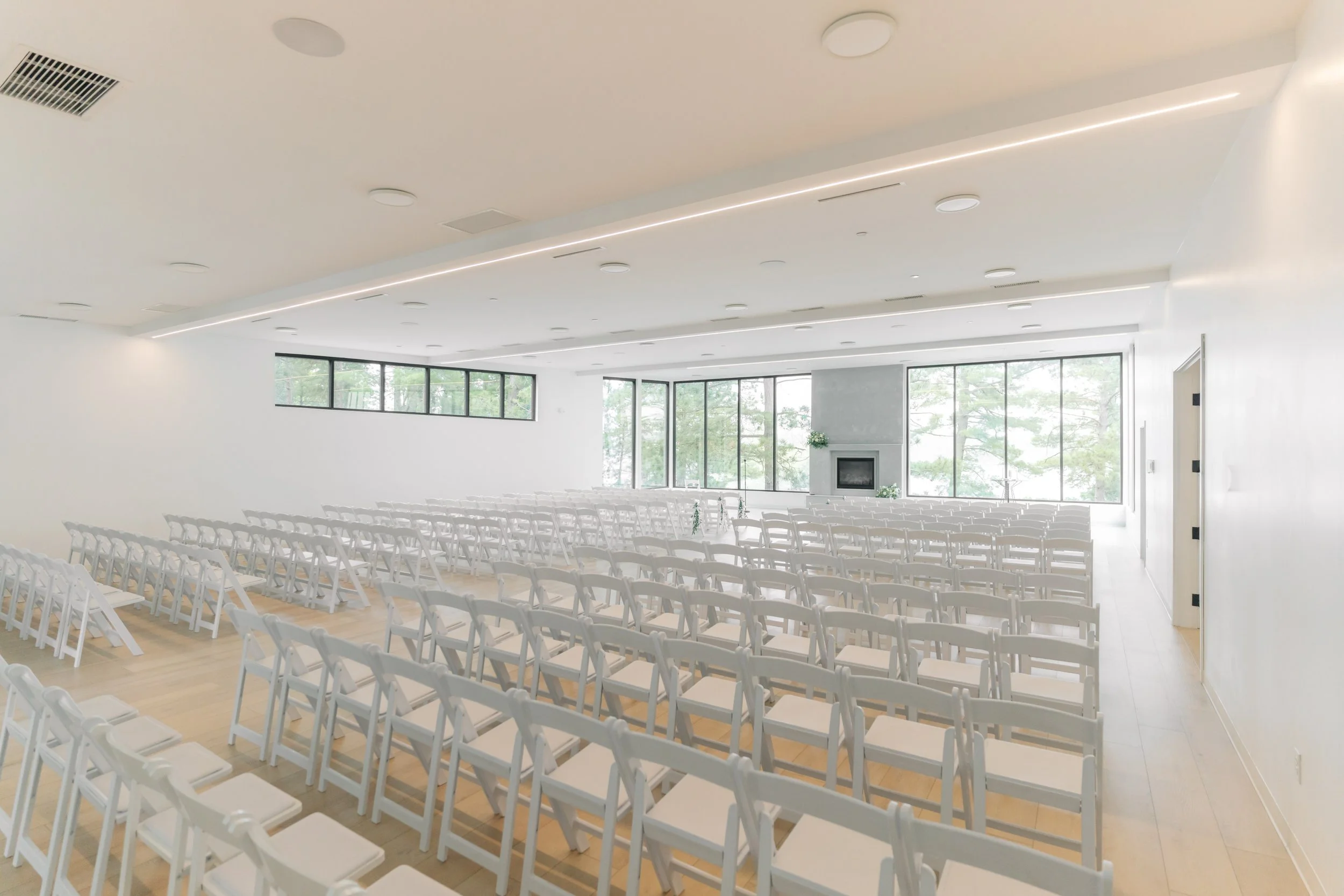 Interior of a modern, minimalistic event space with rows of white chairs facing a large windowed wall, a fireplace, and simple decor.