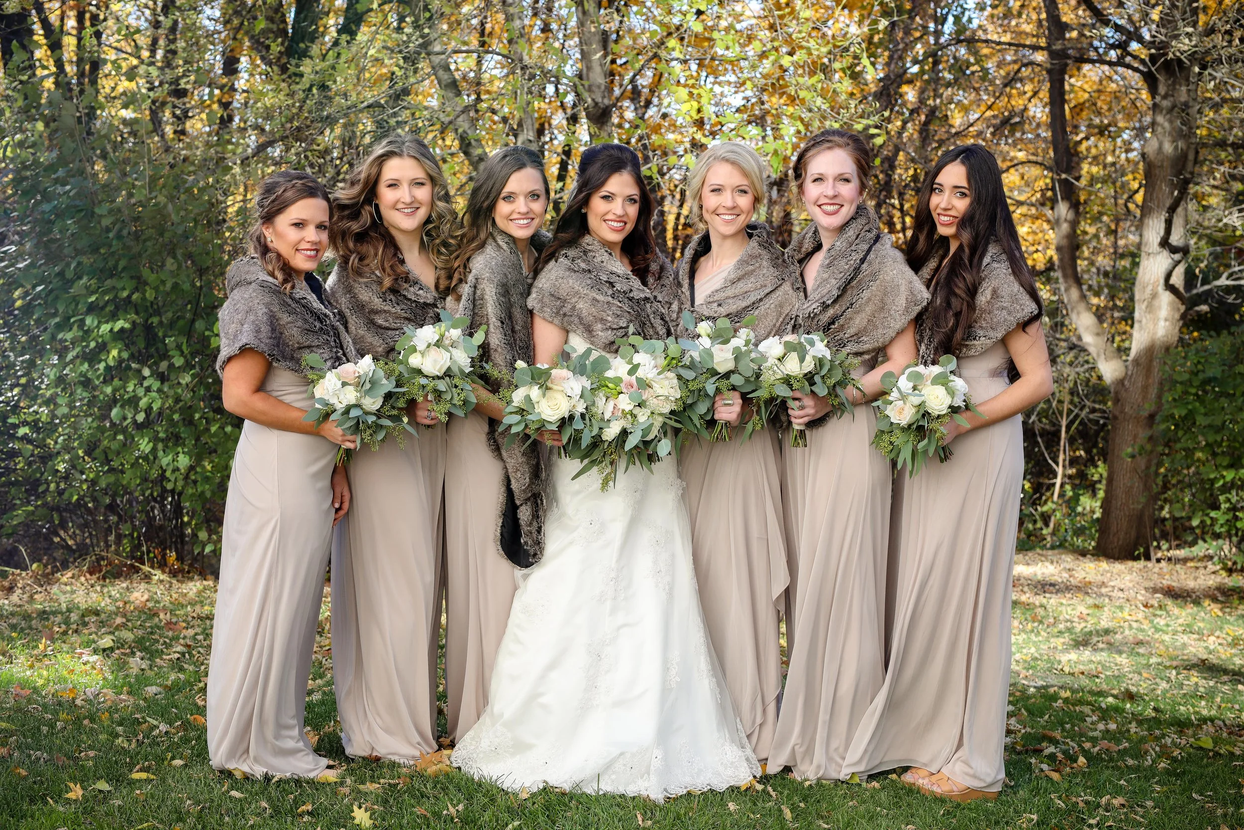 A bride in a white wedding gown holding a bouquet, surrounded by six bridesmaids in beige dresses with fur shawls, holding bouquets, standing outdoors in a forested area with autumn foliage.