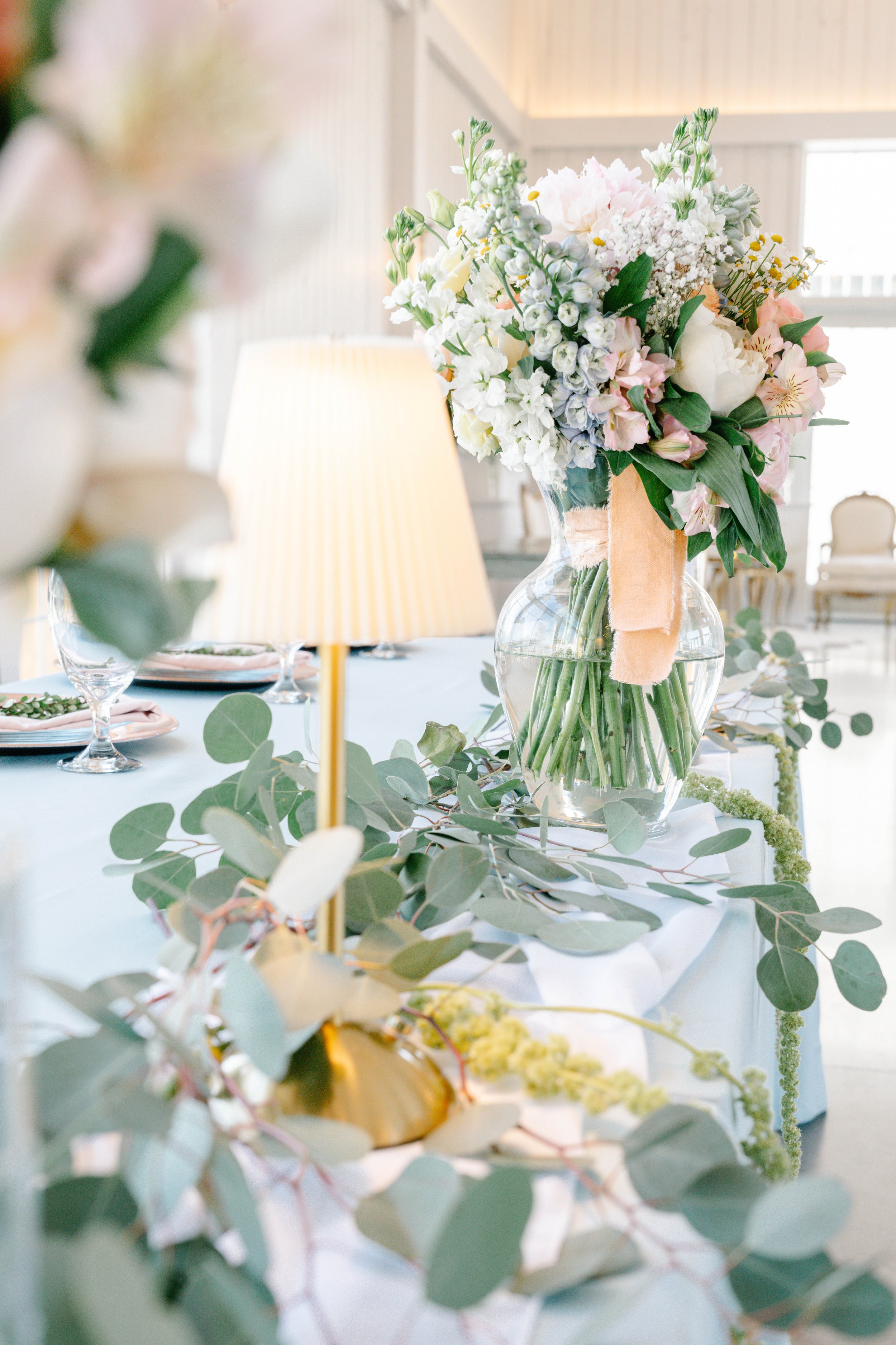 A close-up of a floral centerpiece with white, pink, and purple flowers in a glass vase, on a table decorated with greenery and candles in a bright, elegant room.