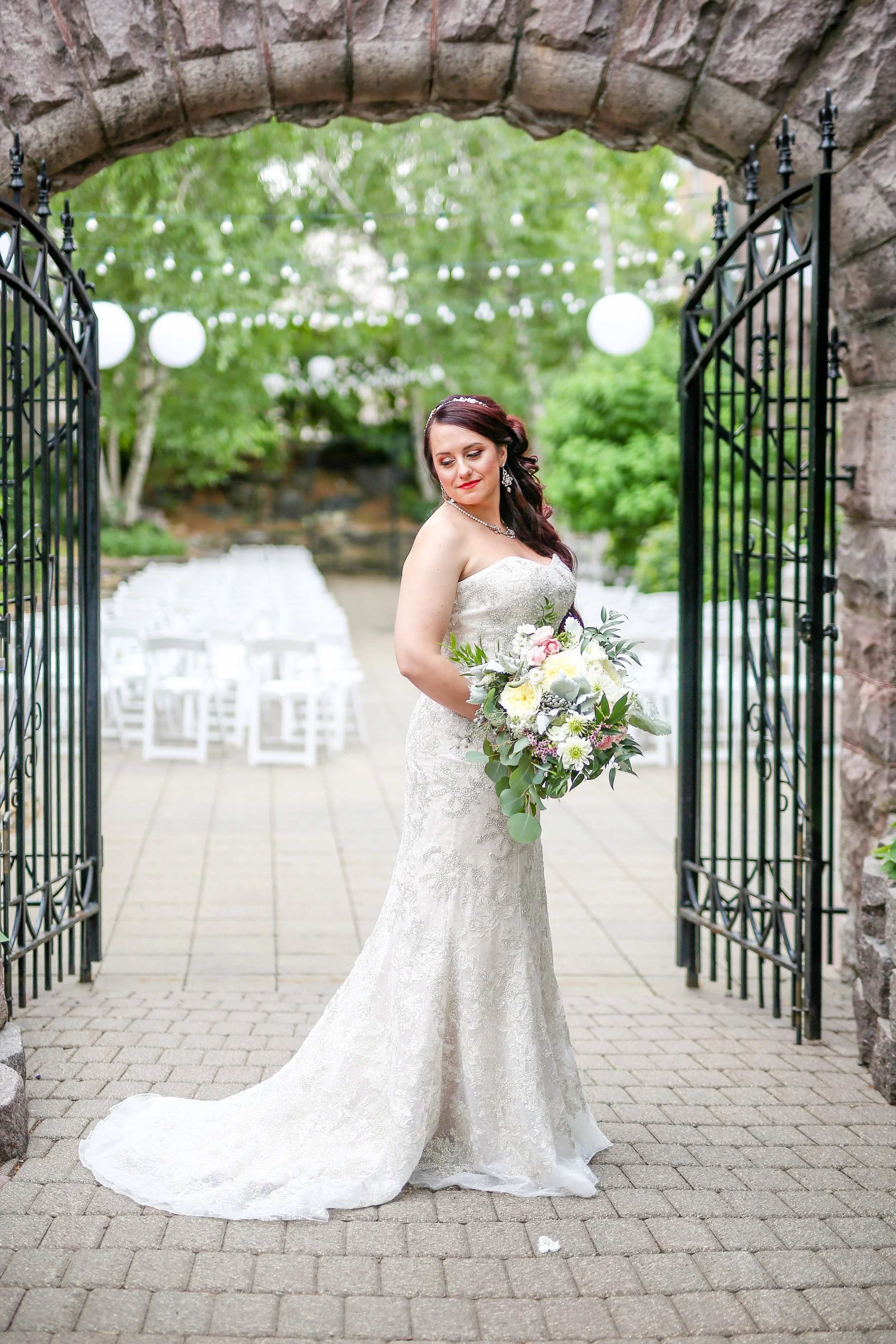 Bride in a white lace wedding dress holding a bouquet of flowers, standing outdoors under a stone archway with greenery and string lights in the background.