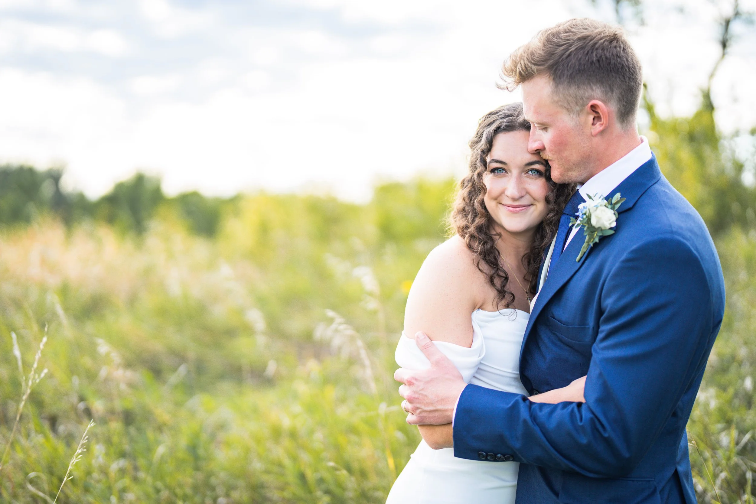 A newlywed couple hugging outdoors in a field of tall grass and trees, the bride in a white dress with dark curly hair, and the groom in a blue suit with a boutonniere, taken by Minnesota Wedding Photographer, LKW Photography.