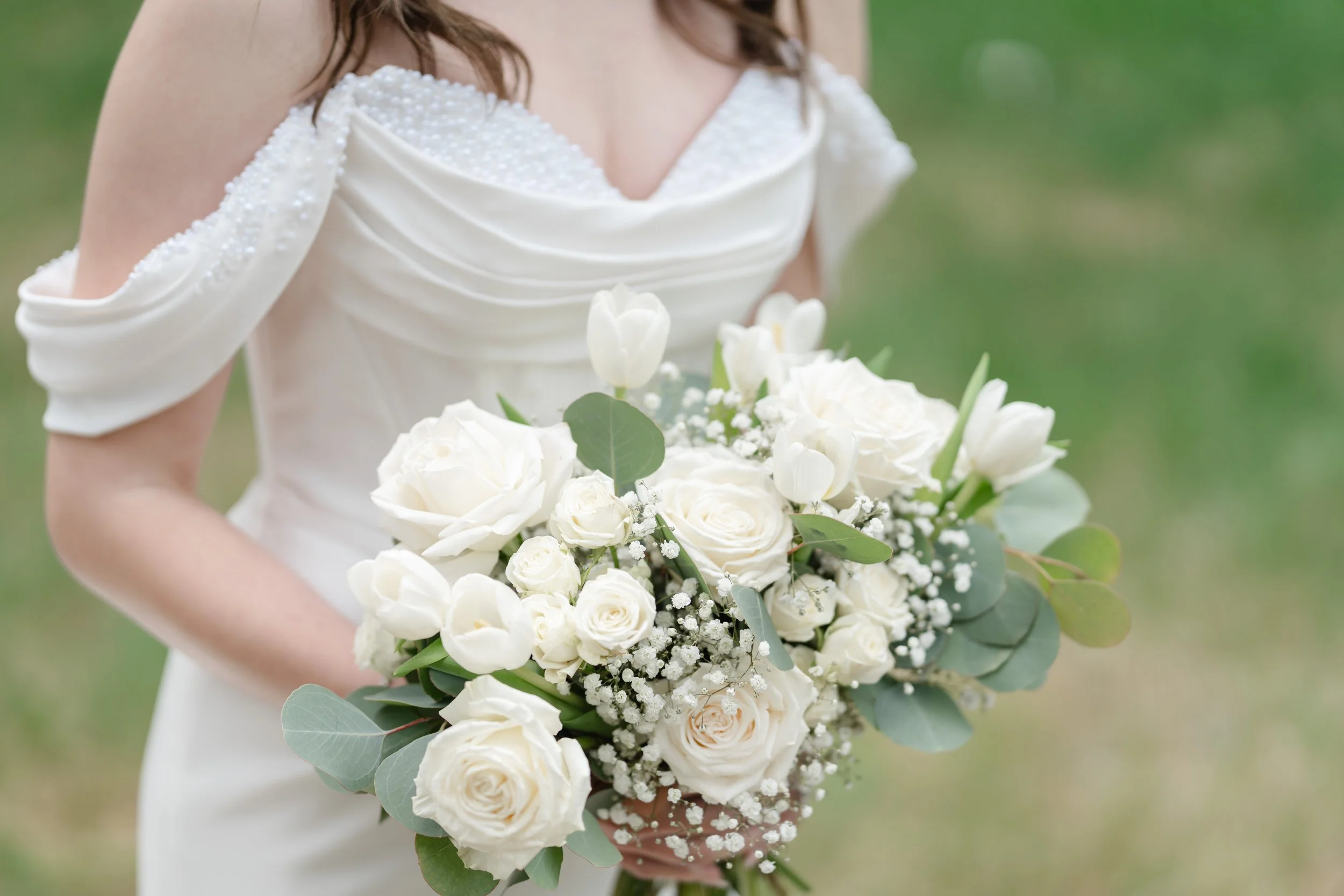 A bride holding a bouquet of white roses, tulips, and eucalyptus leaves, wearing a white off-shoulder wedding dress with pearl embellishments on the shoulder.