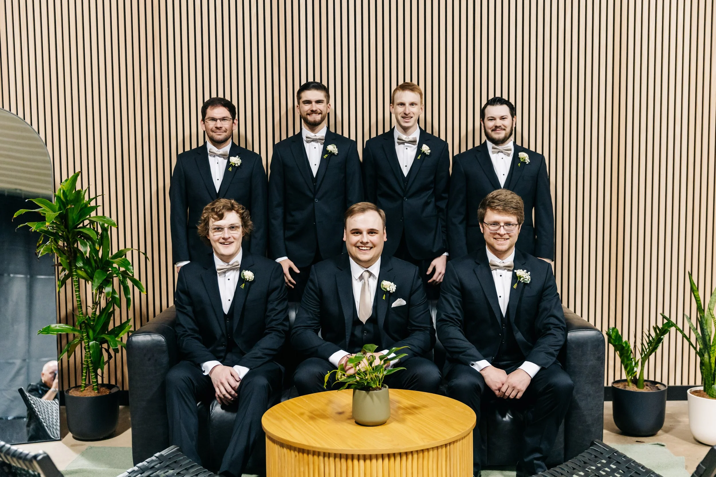 Six men dressed in formal tuxedos with bow ties and boutonnières, posing for a group photo in front of a wood-paneled wall, with potted plants and round wooden table in the foreground.