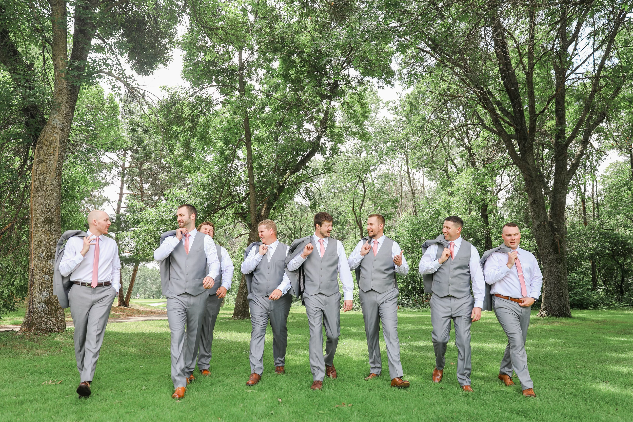 A group of men in gray suits and pink ties walking together outdoors in a park with green grass and trees, carrying jackets over their shoulders, by LKW Photography.
