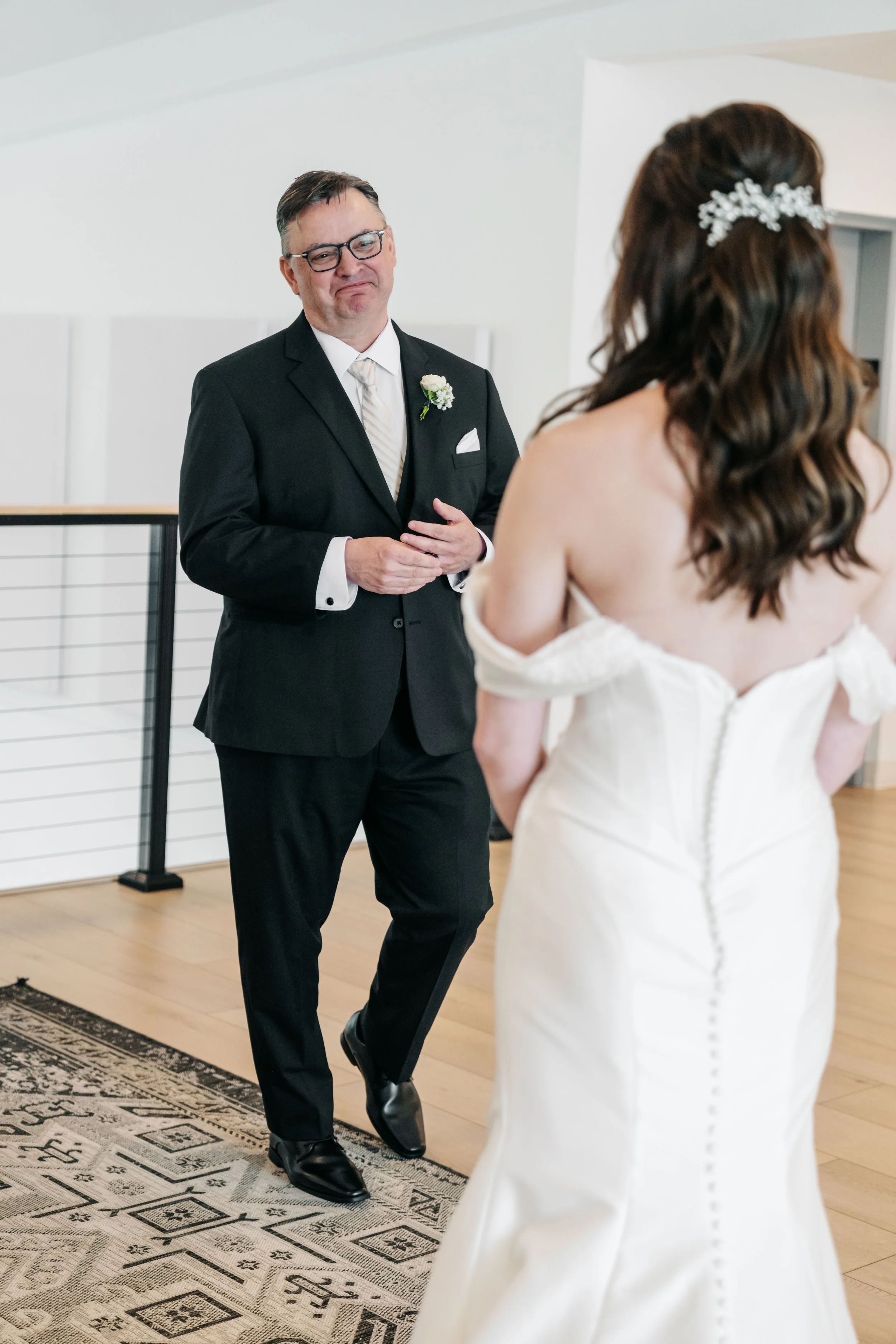 A man in a black suit and glasses is smiling looking at a woman in a white wedding dress with long brown hair and a floral hair accessory. They are indoors, standing on a patterned rug.