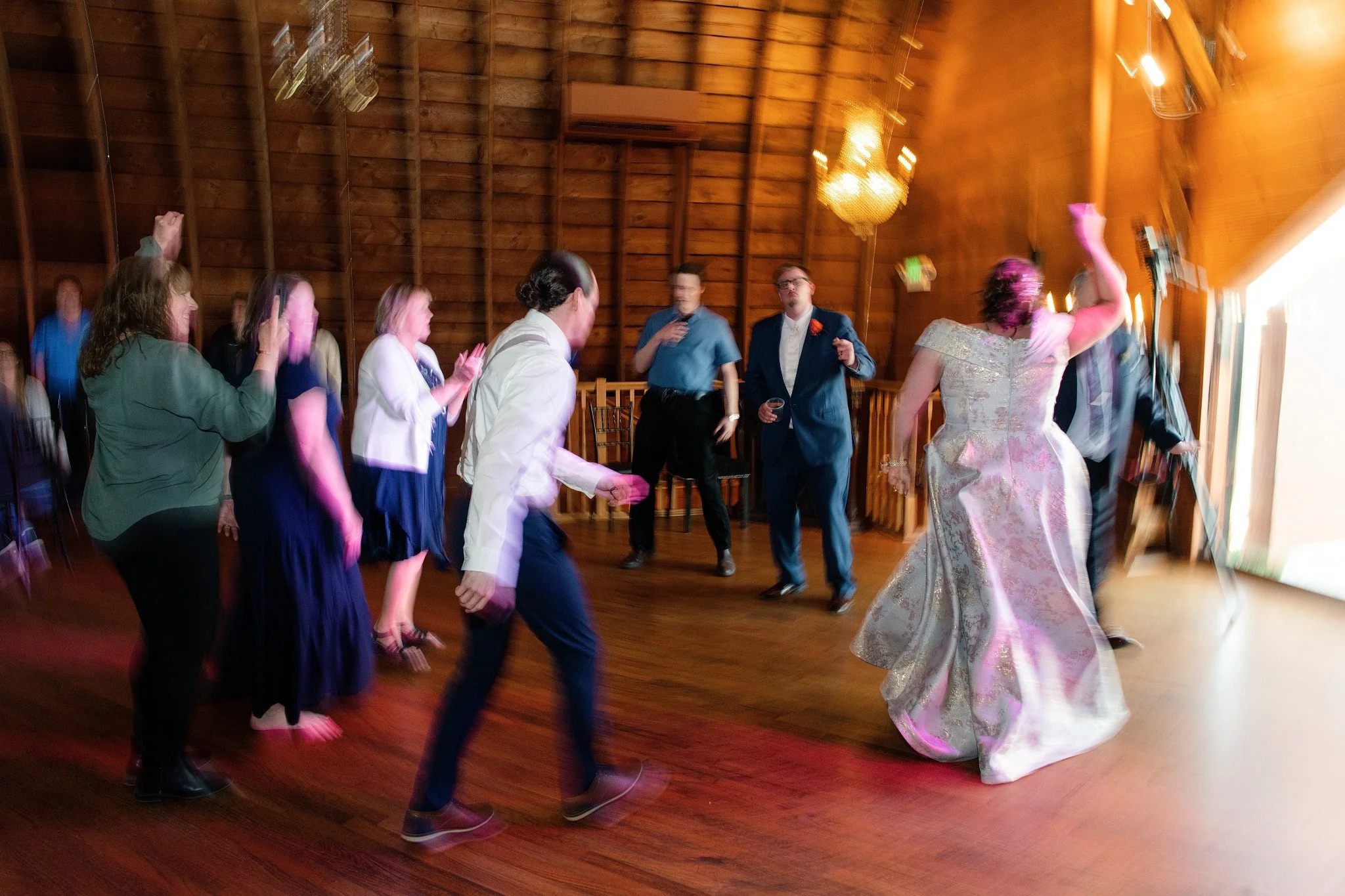 People dancing at a celebration or wedding reception inside a wooden venue with a chandelier, including a woman in an elegant gown, and others in formal attire, moving to music.
