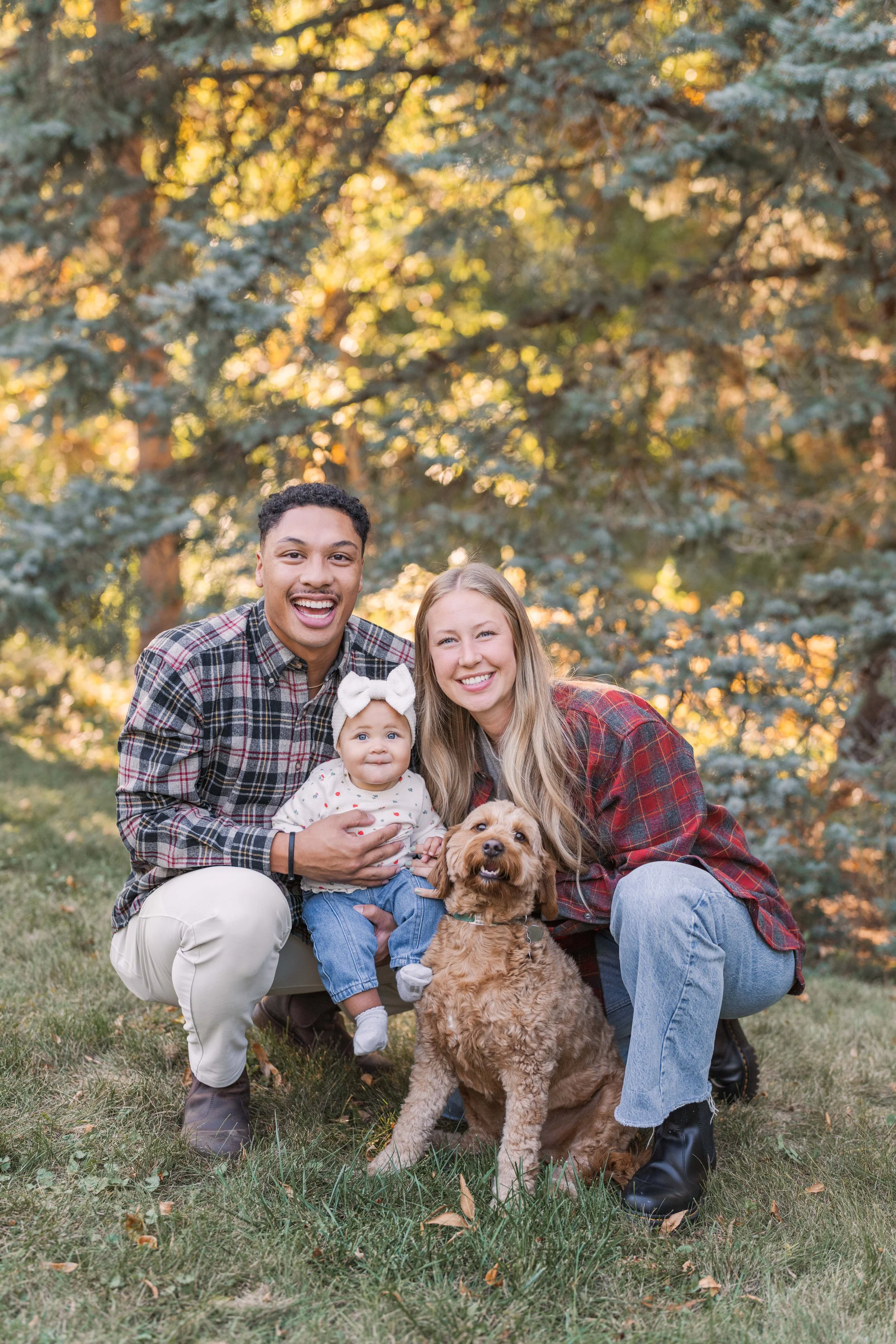 Minnesota portrait photographer, LKW Photography, photographs smiling family of four, including a man, woman, a baby girl with a white bow, and a dog, outdoors in a backyard with trees.
