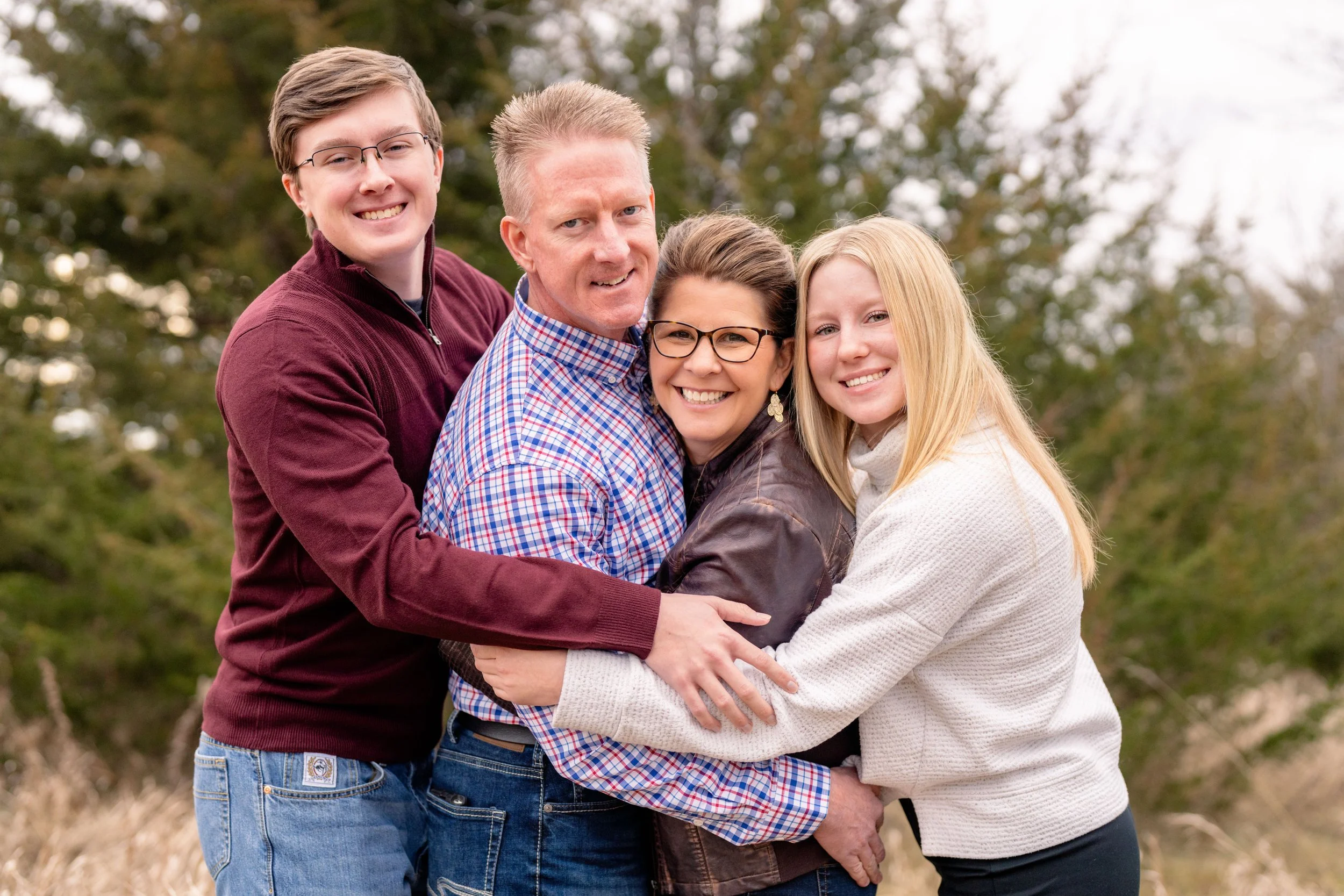 Minnesota family portrait photographer, LKW Photography, photographs a family of four hugging outdoors with trees in the background, smiling at the camera.