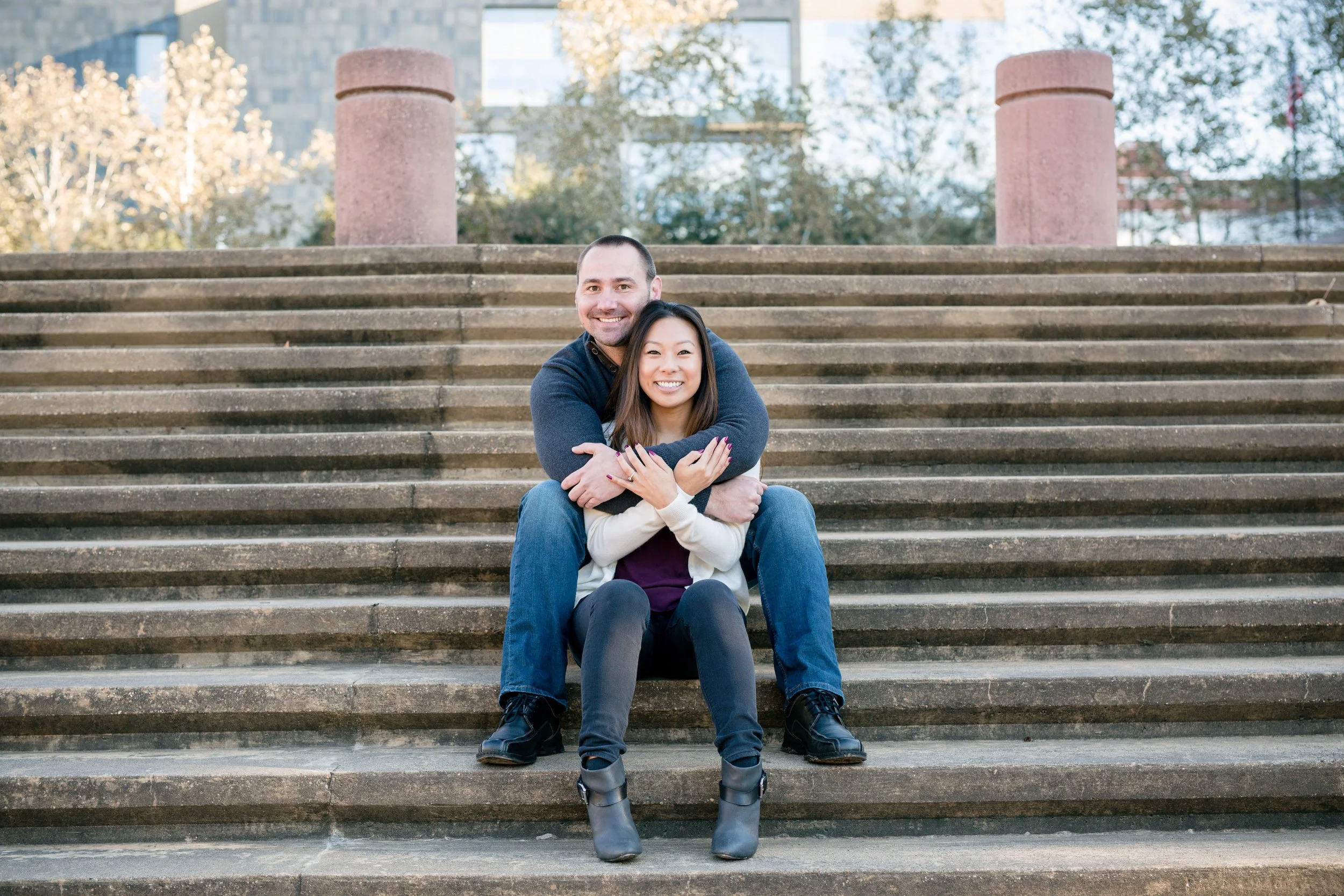 A smiling man and woman sitting on outdoor stairs with the man hugging the woman from behind. Trees and buildings are in the background, taken by MN photographer LKW Photography in Austin, TX.