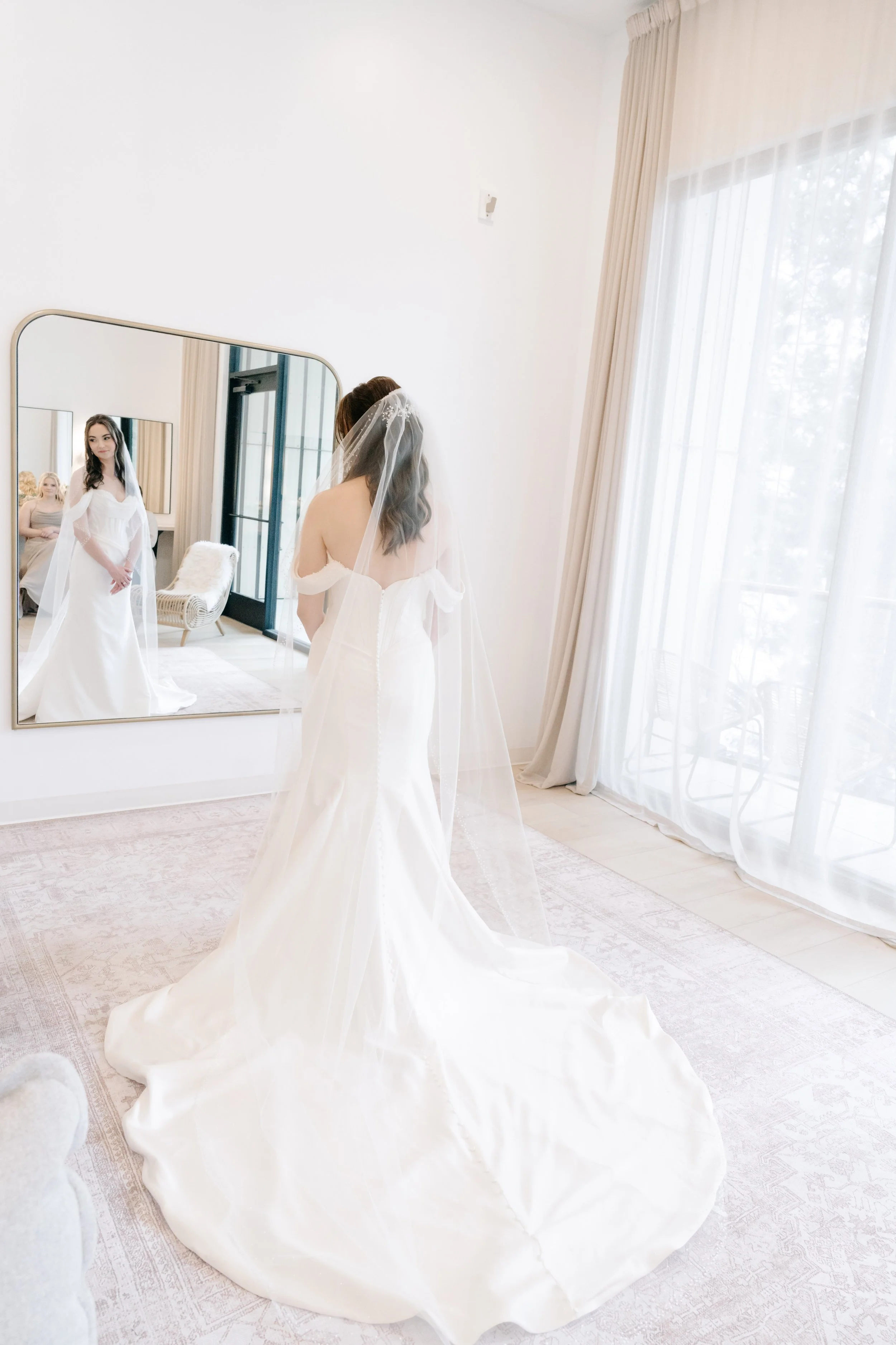A bride in a white wedding dress and veil looking at herself in a large mirror in a bright room with light-colored curtains and furniture.
