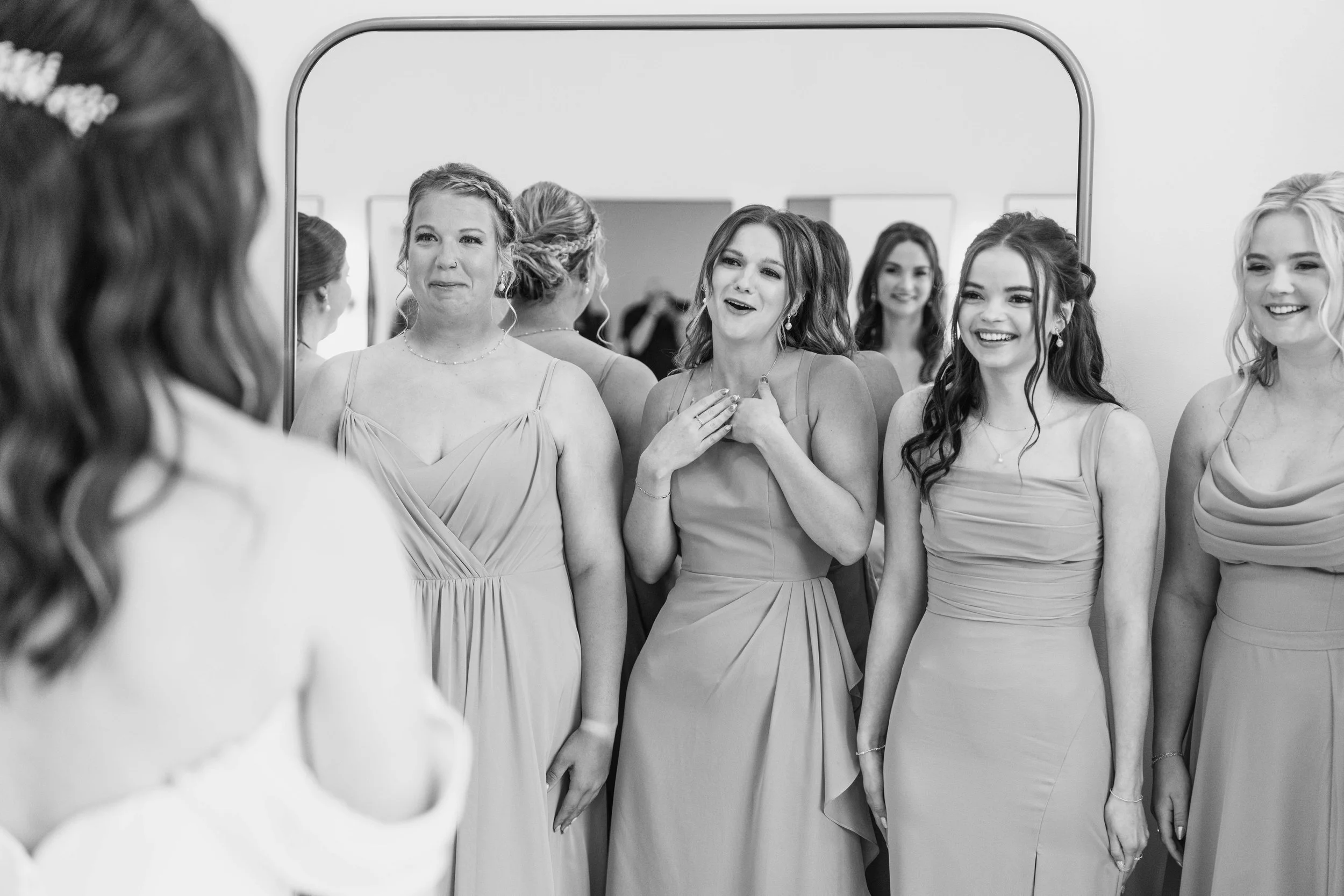 A group of women in formal dresses looking at themselves in a mirror, showing various expressions of happiness and surprise.