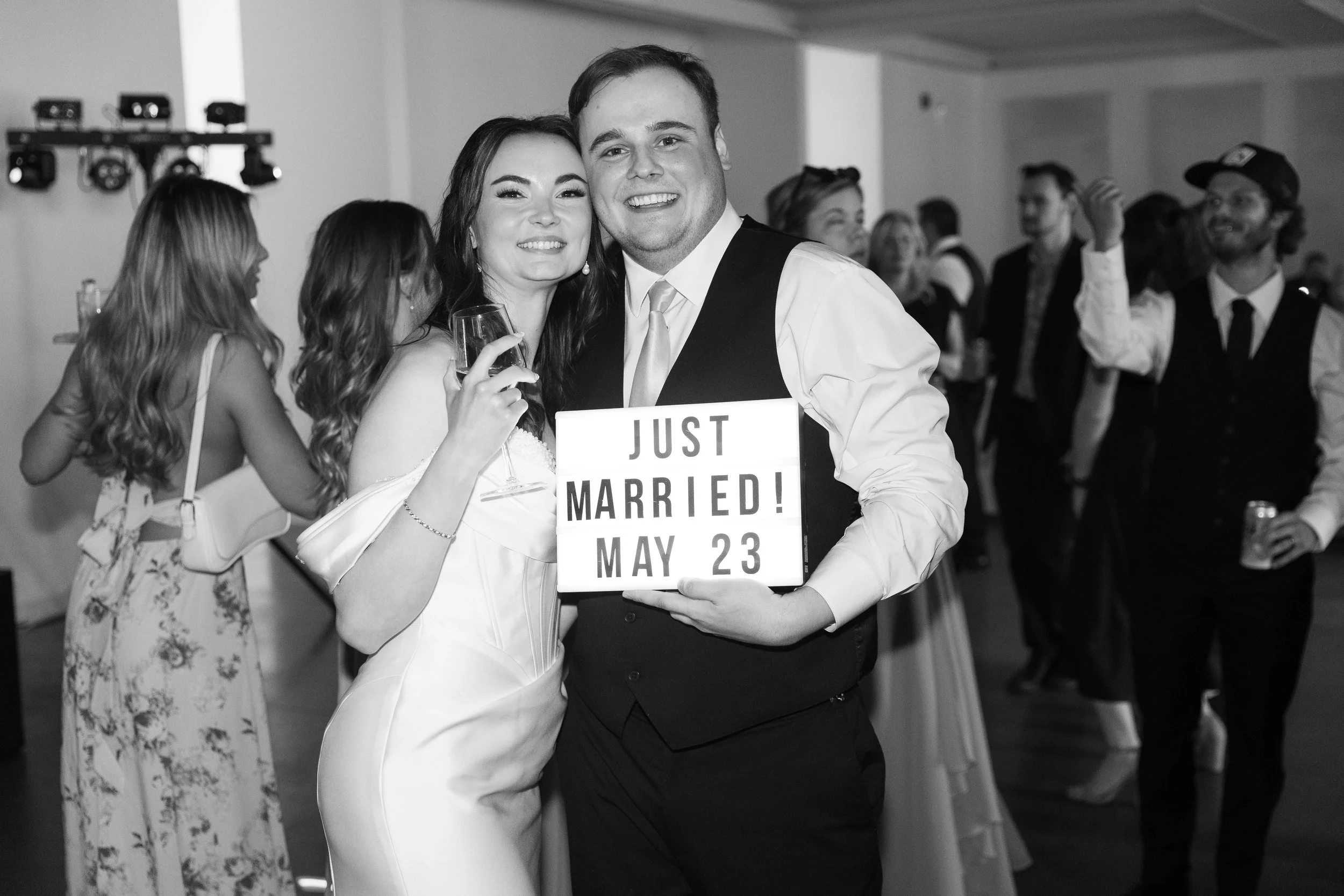 A smiling bride and groom at their wedding reception, holding a 'Just Married' sign with their wedding date, May 23. The bride is dressed in a wedding gown, and the groom is in a vest and tie. They are surrounded by guests in a decorated indoor venue