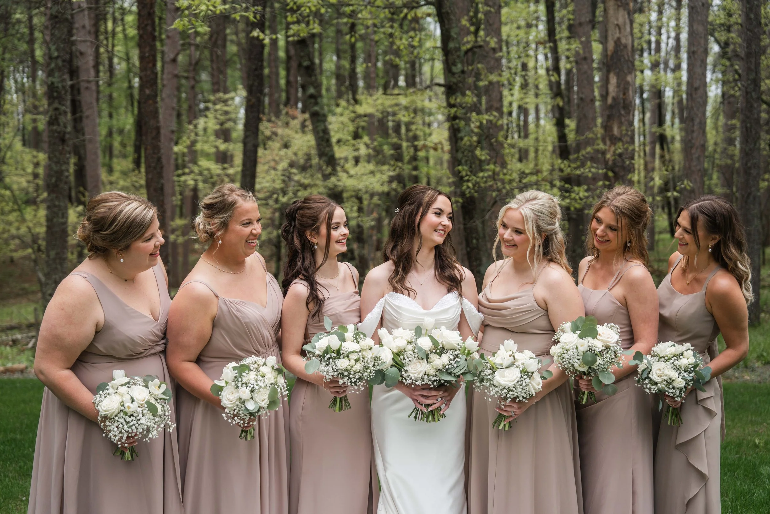 A bride and six bridesmaids stand outdoors in a wooded area, smiling and holding bouquets of white flowers and greenery.