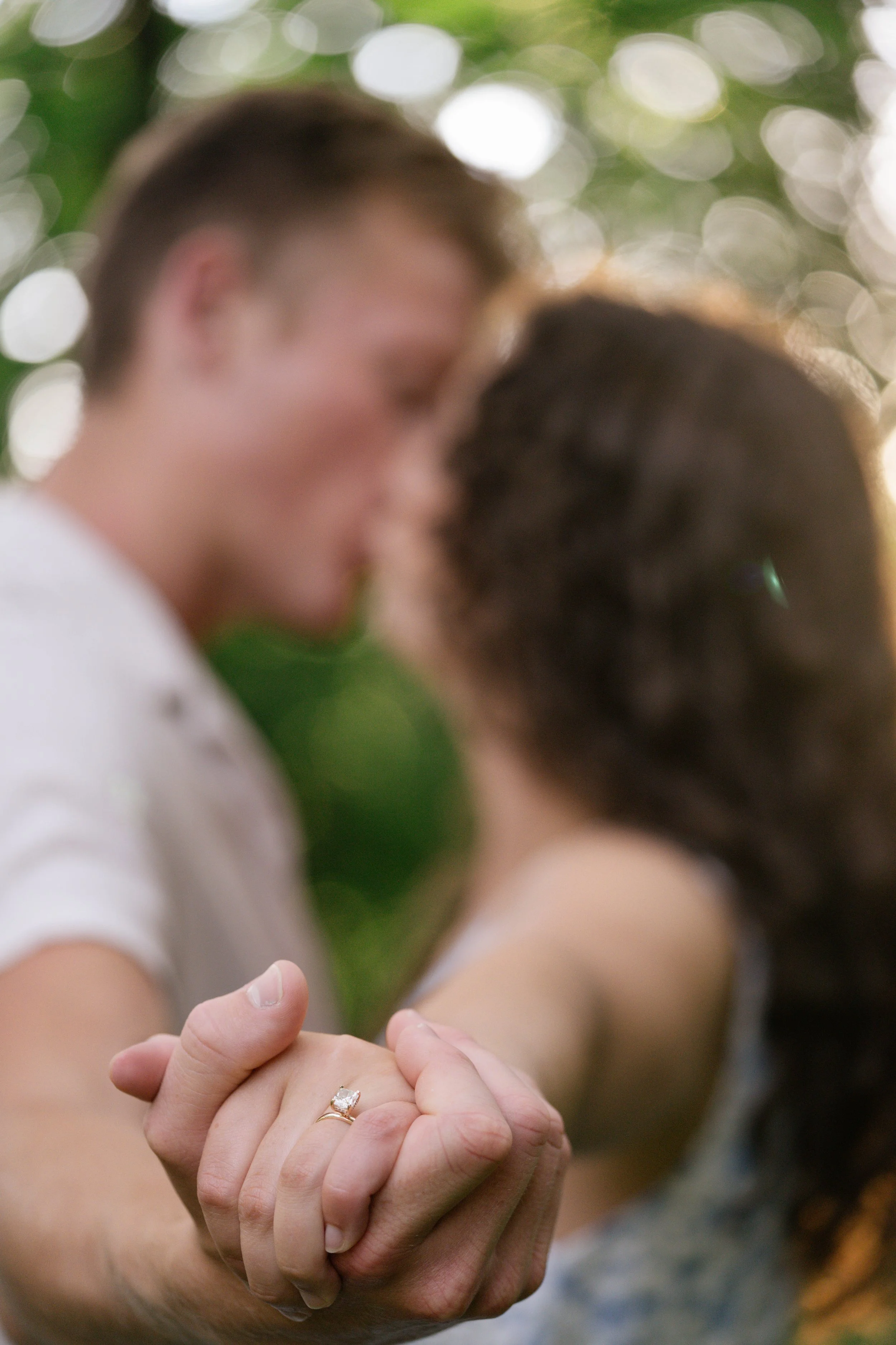 An engagement session with the couple holding hands outdoors with a focus on the woman's hand showing an engagement ring, while their faces are blurred in the background, taken by MN photographer LKW Photography.
