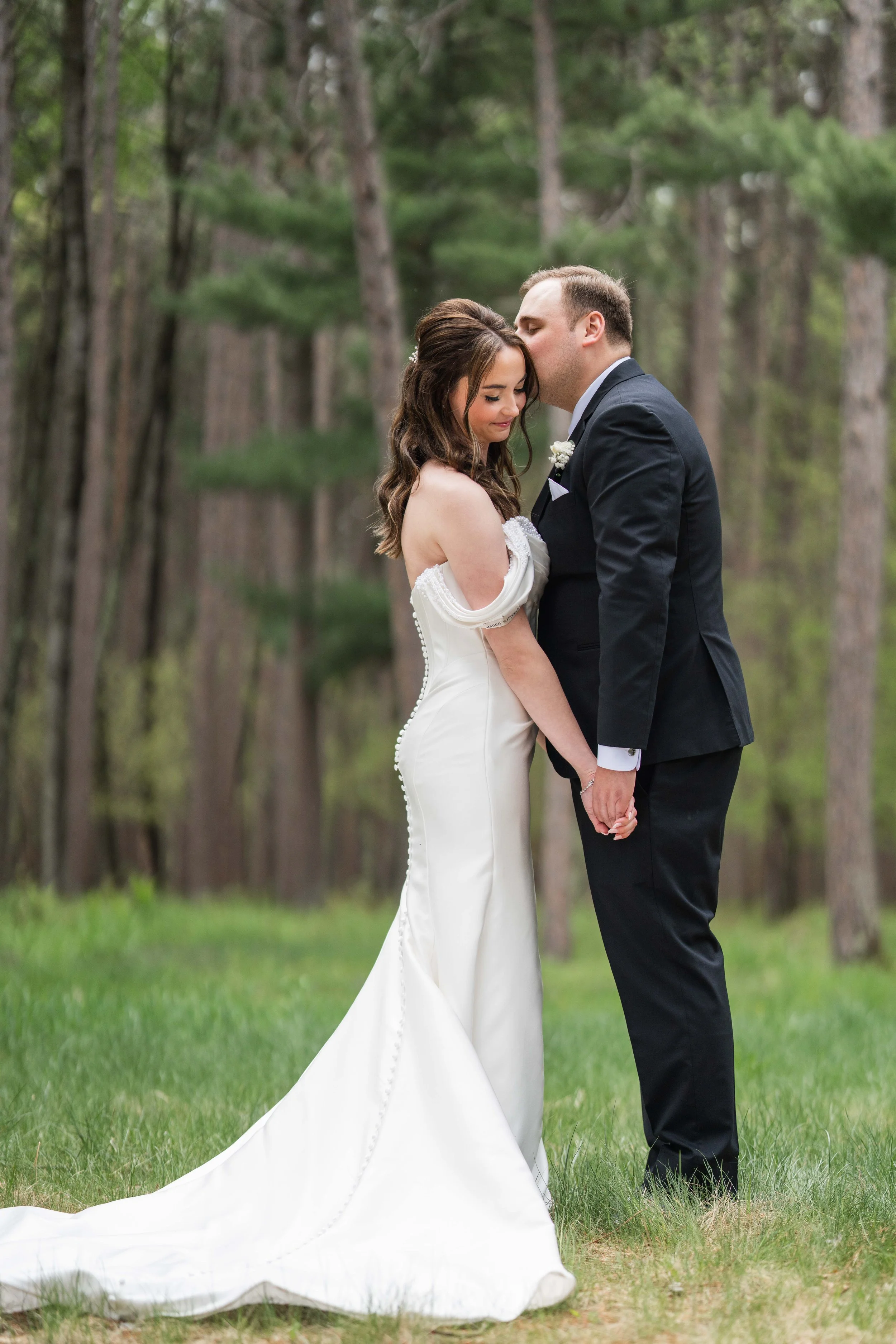 A bride and groom stand close together in a forest, with the groom kissingly touching the bride's forehead while she gently holds his hand, both with their eyes closed, sharing an intimate moment during their wedding.