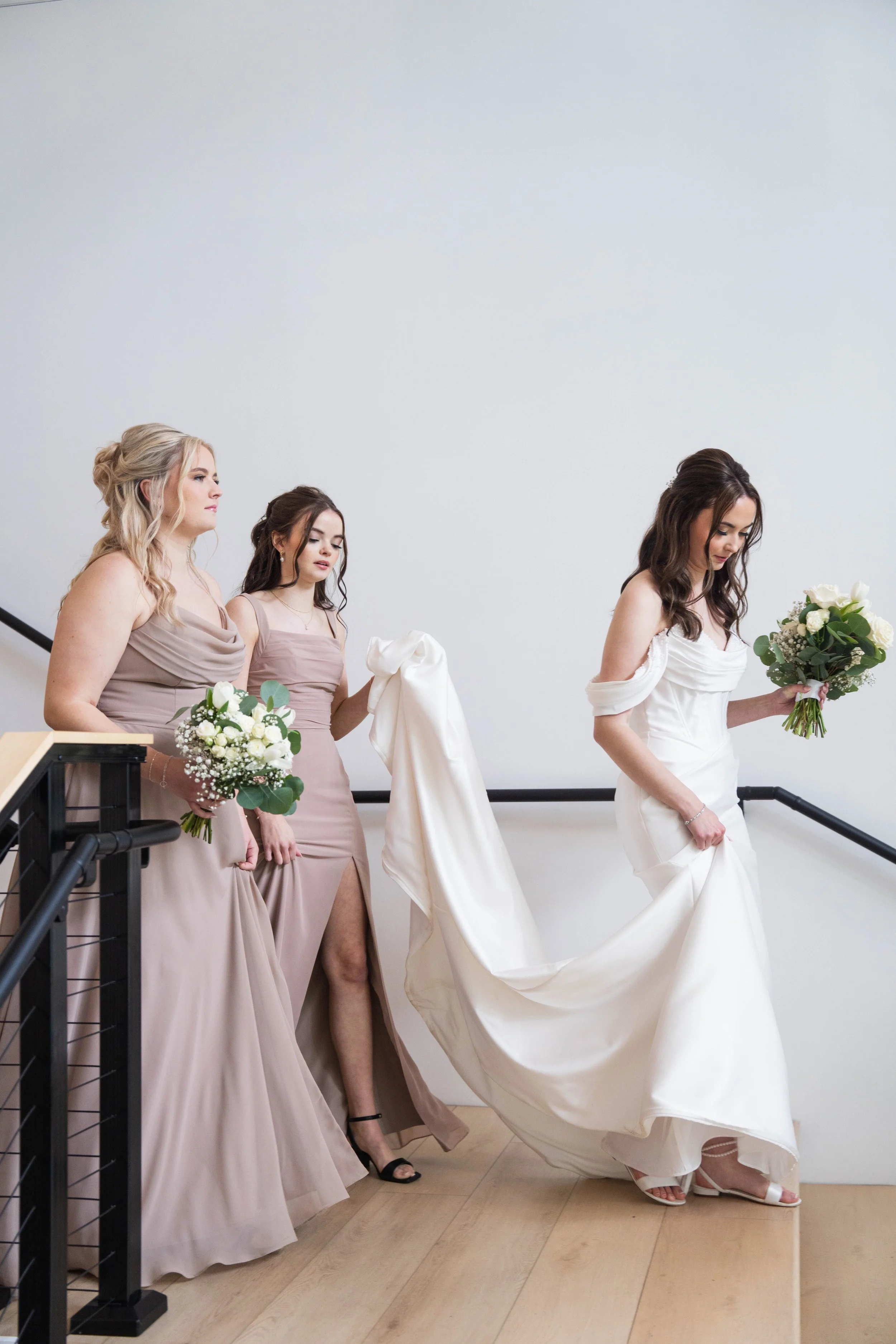 Three women in formal dresses holding bouquets, standing on a staircase against a plain wall, with one woman adjusting her dress and the others looking on.