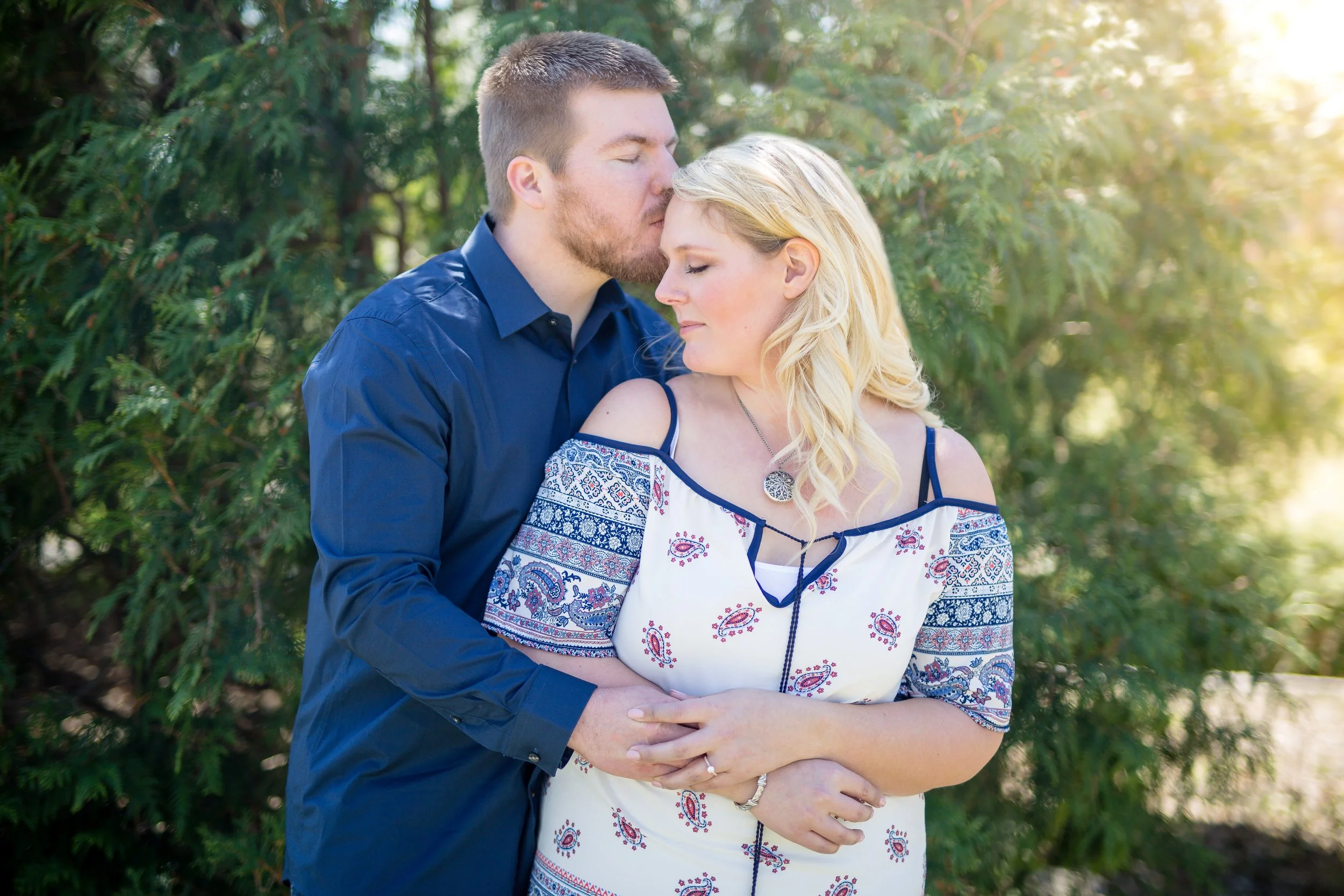 A couple embraces outdoors in front of green trees, with the man kissing the woman's forehead as she closes her eyes and smiles, taken by Minnesota wedding photographer, LKW Photography.