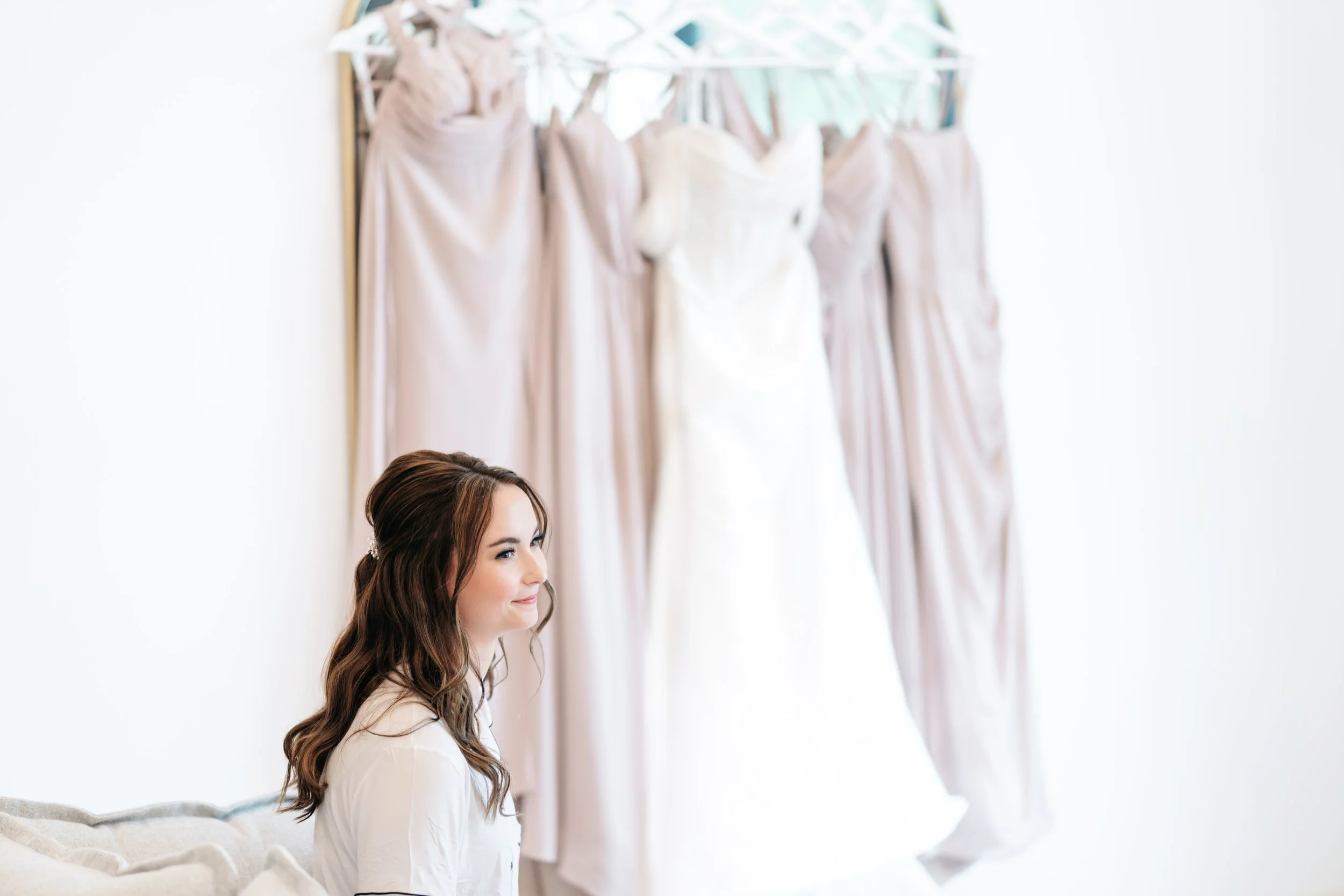 A woman with long brown hair and a white top is sitting indoors near a white wall with dresses hanging in the background.
