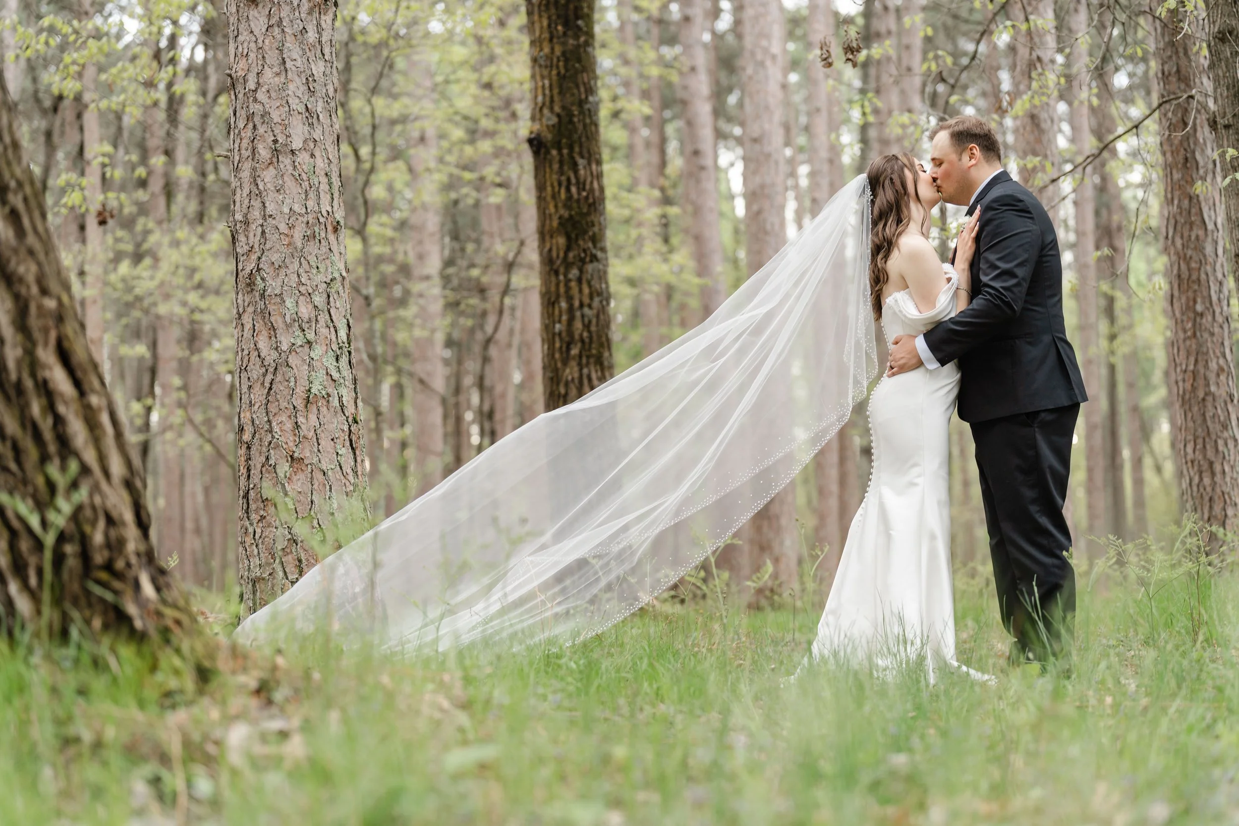 A bride and groom sharing a kiss in a wooded forest, with the bride's long veil flowing behind her.