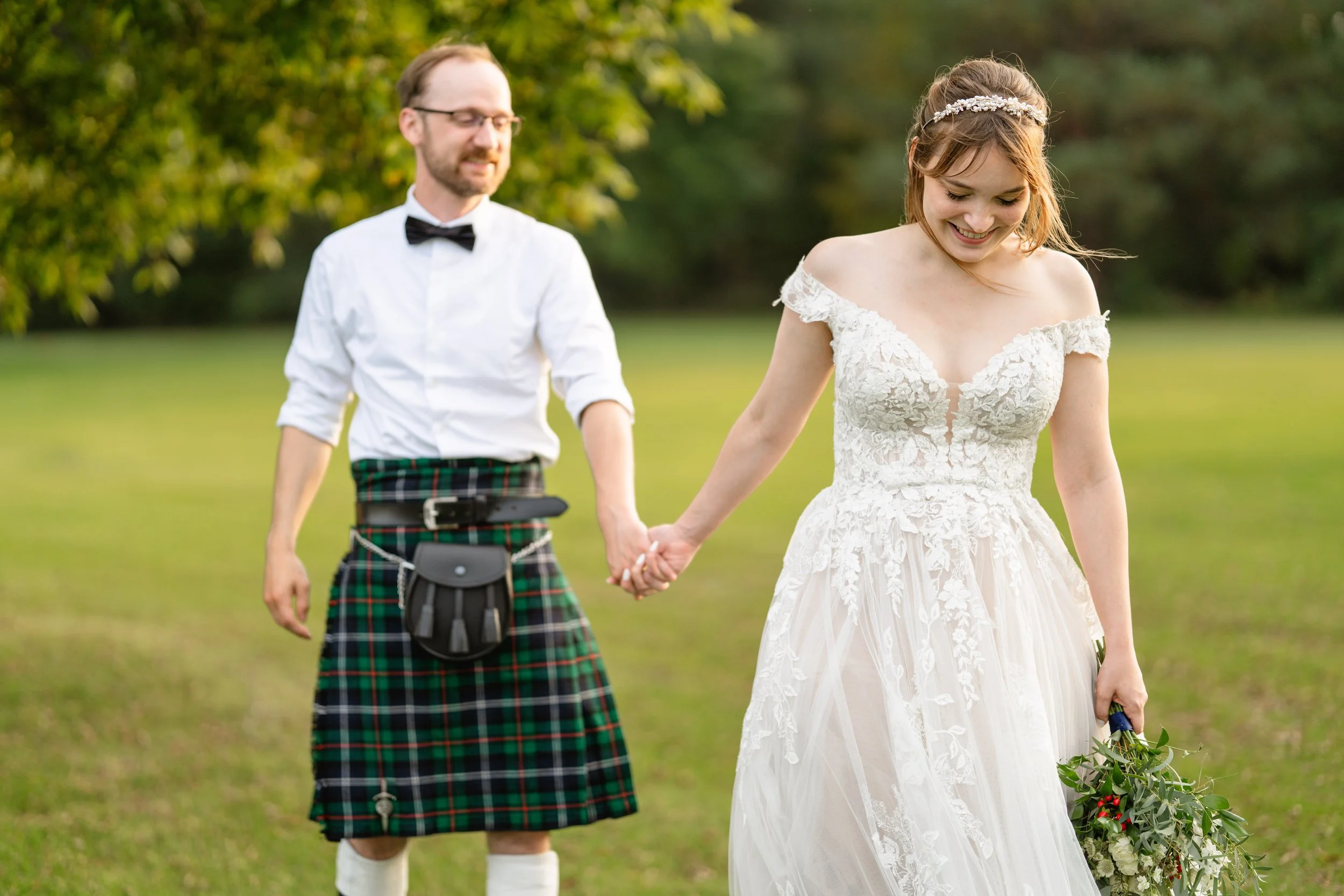 A bride in a white lace wedding dress holding a bouquet, walking outdoors with a groom in a traditional Scottish kilt, on a grassy field with trees in the background.