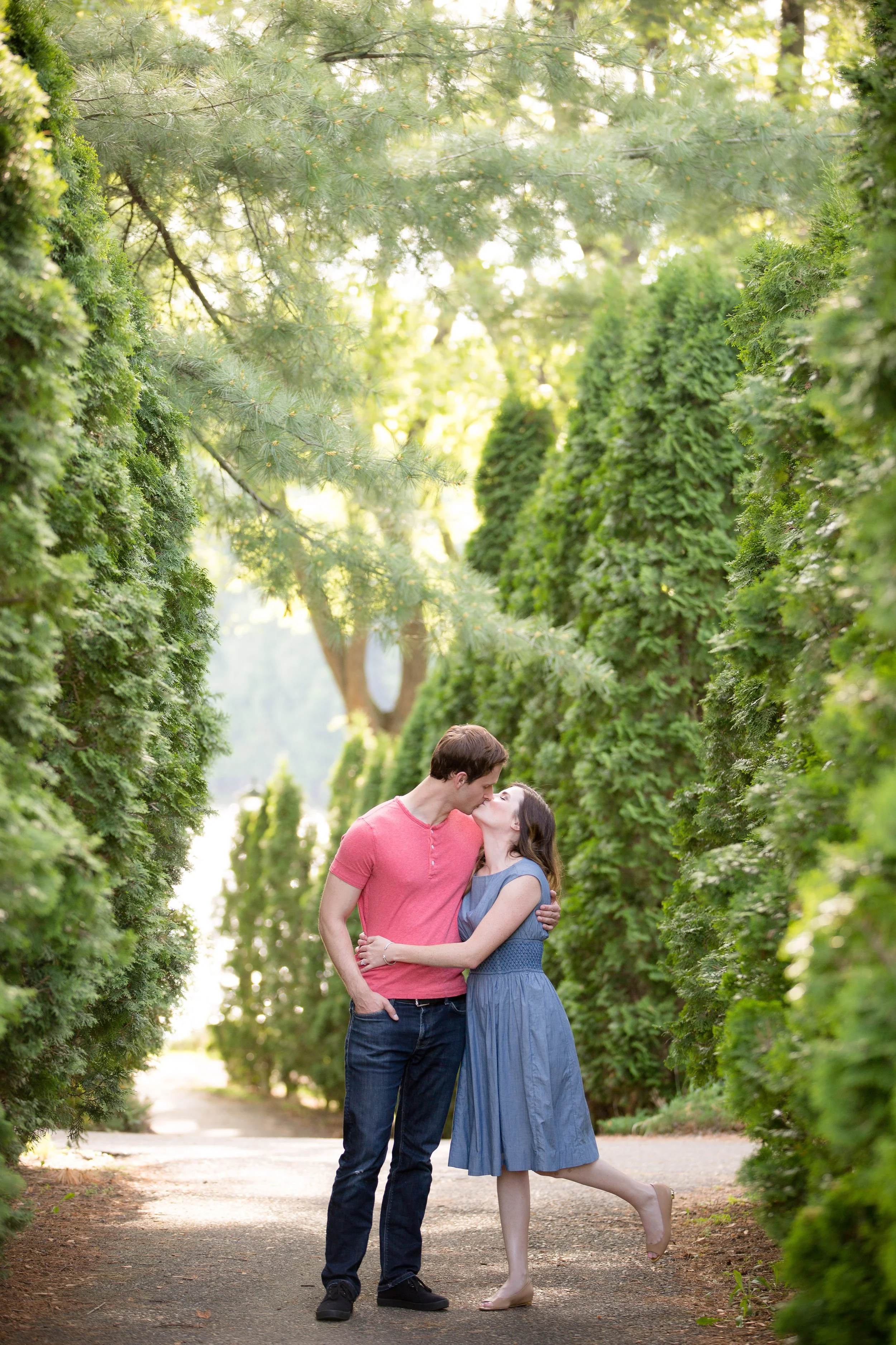 An engaged couple kissing in a lush green park with tall trimmed bushes and trees, taken by MN photographer LKW Photography in St. Cloud.