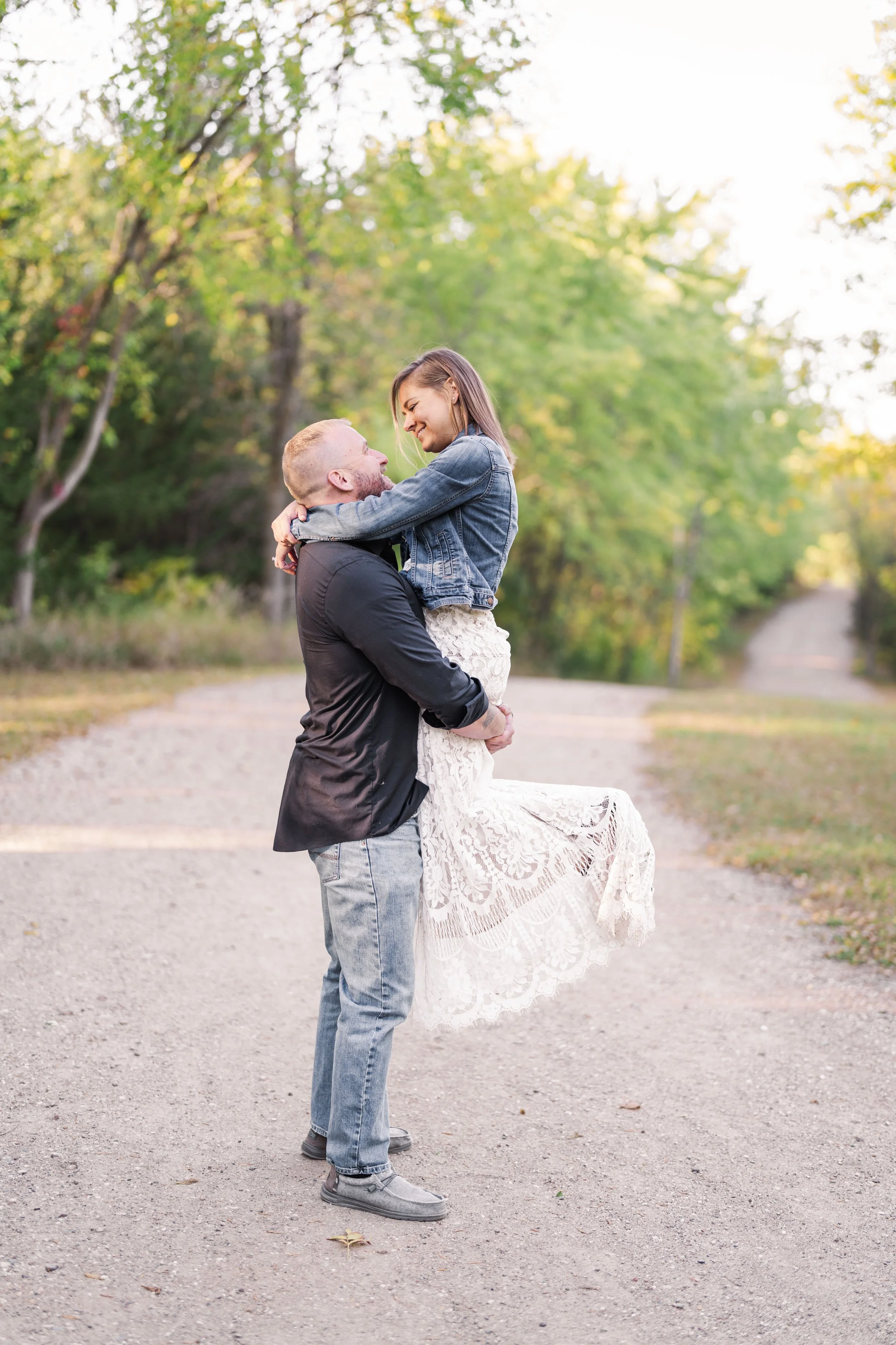 Minnesota wedding photographer, LKW Photography, photographs an engagement session with a man lifting a woman in an outdoor setting with trees in the background, both smiling and looking at each other.